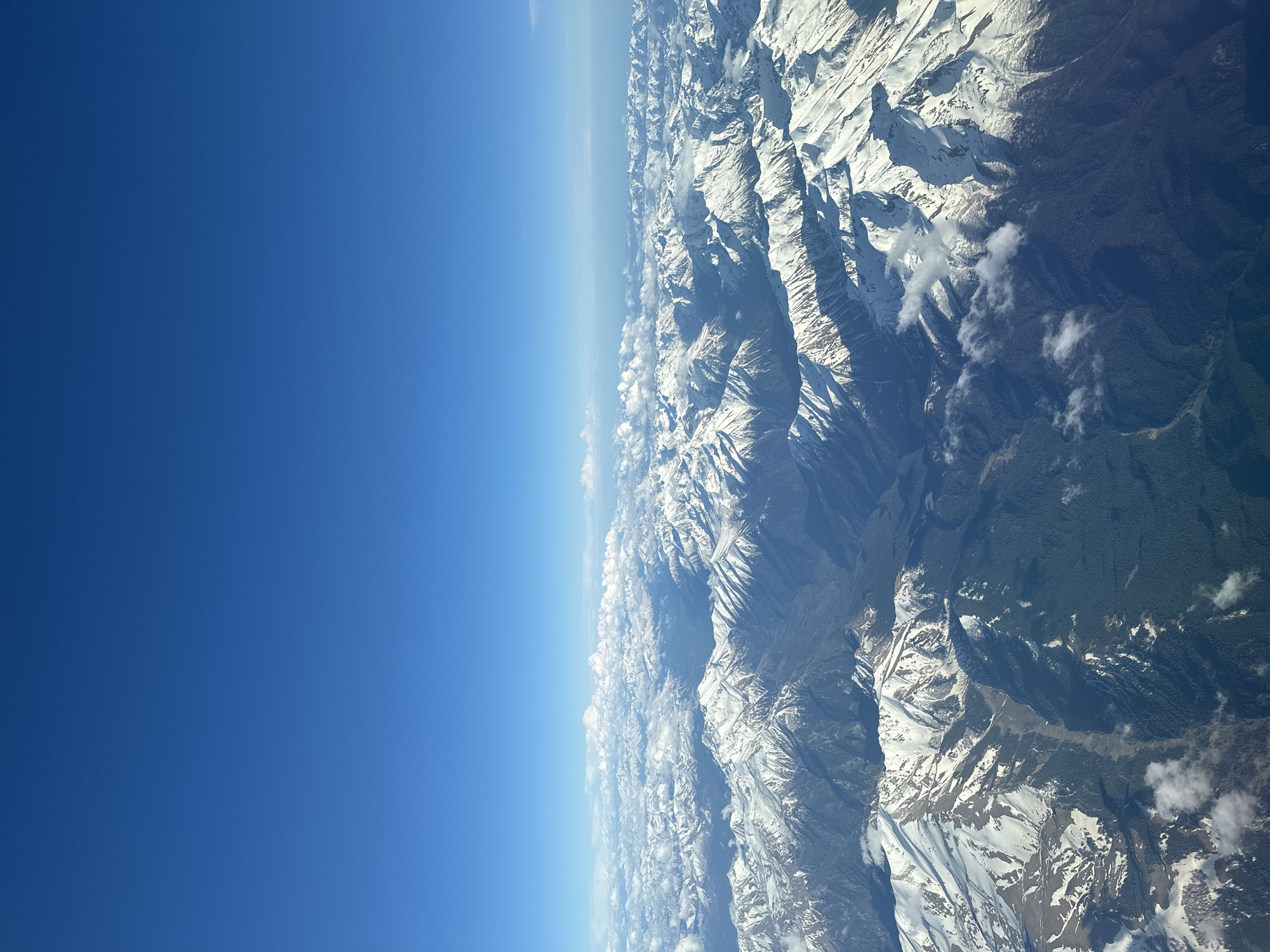 The image shows a view of mountains covered in snow, likely taken from an airplane or high altitude. The left side of the image is dominated by a clear, blue sky. The mountains are rugged and textured, with areas of exposed rock and snow-covered peaks. Clouds are interspersed among the mountains. There is no visible location in the city of Khimki, Russia, people, subjects, objects, actions, or interactions visible in the image.