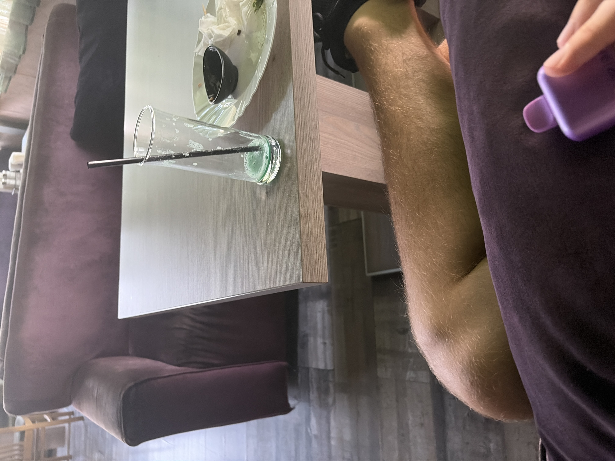 The image captures a close-up view of a person's bare leg resting on a dark purple fabric, likely pants or a blanket. A small, purple electronic vape device is held in the person's hand, which is partially visible in the lower right corner. In the background, a wooden table holds a used glass with a black straw and a small black dish on a plate, suggesting a meal or drinks were consumed. The setting appears to be indoors, possibly a cafe or restaurant, with a plush purple sofa visible in the upper left quadrant. The floor is a wood-like laminate. The scene does not provide specific information to pinpoint its location in Elin Pelin, Bulgaria, beyond it being an indoor establishment.