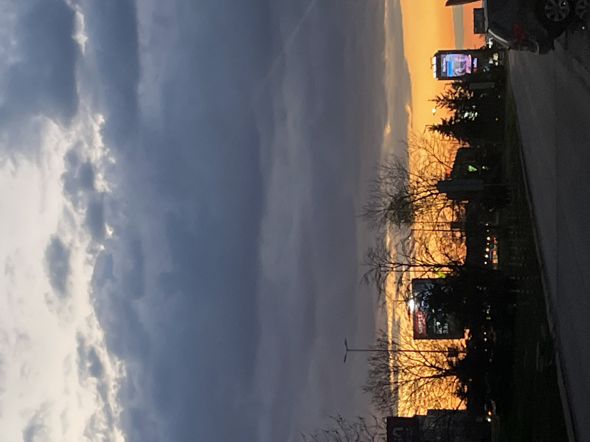 The image depicts a sunset in an urban setting, likely Sofia, Bulgaria. The sky on the left side is dominated by dark, heavy clouds, while the right side displays a vibrant orange and yellow sunset. Silhouetted against the sunset are bare trees, street lights, and buildings. There is a large billboard illuminated with a bright screen. A road runs along the right side of the frame with a car parked on the side.