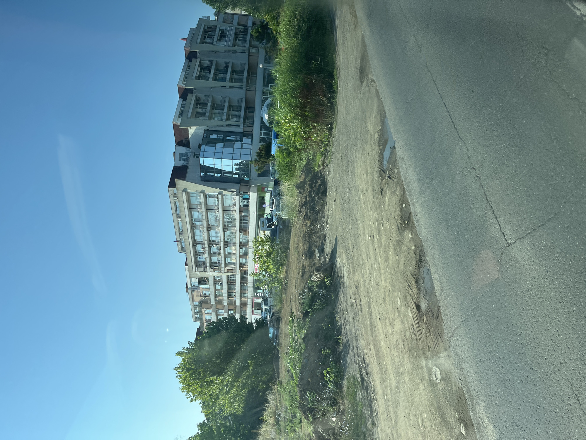 The image shows a multi-story building with a modern architectural design, featuring balconies and large glass windows, located in the city of Nesebar, Bulgaria. In front of the building, there is a plot of undeveloped land with dirt mounds and sparse vegetation. Several vehicles, including cars and what appears to be a van, are parked in the vicinity of the building. The foreground of the image displays a paved road with some visible cracks and a small puddle of water. The sky is clear and blue, suggesting a sunny day.