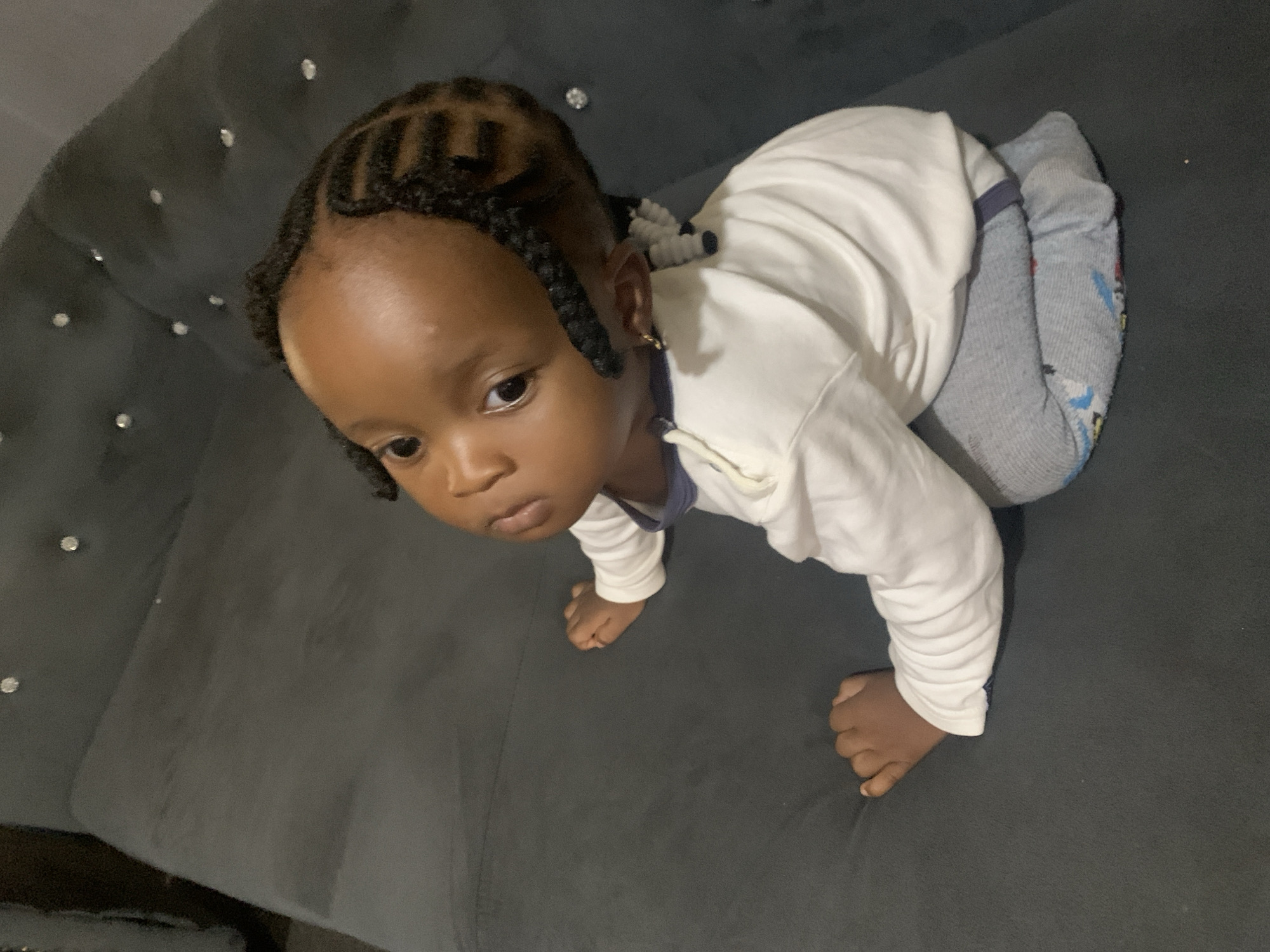 The media file shows a close-up, high-angle shot of a young Black child crawling on a dark gray tufted couch. The child has dark skin and is wearing a white long-sleeved shirt and gray socks with a cartoon pattern. The child's hair is styled in small braids. The child's face is turned towards the camera, with a neutral expression. The lighting suggests an indoor setting, possibly during the day due to the overall brightness, but the specific time of day is not discernible. The couch appears to be upholstered in a fabric with a subtle sheen and is adorned with small, sparkly embellishments. The background is predominantly the dark gray of the couch, with no other objects or people visible. There is no visible text in the image. The child's posture and the way they are looking suggest they are actively exploring or moving. The overall mood is calm and domestic.
