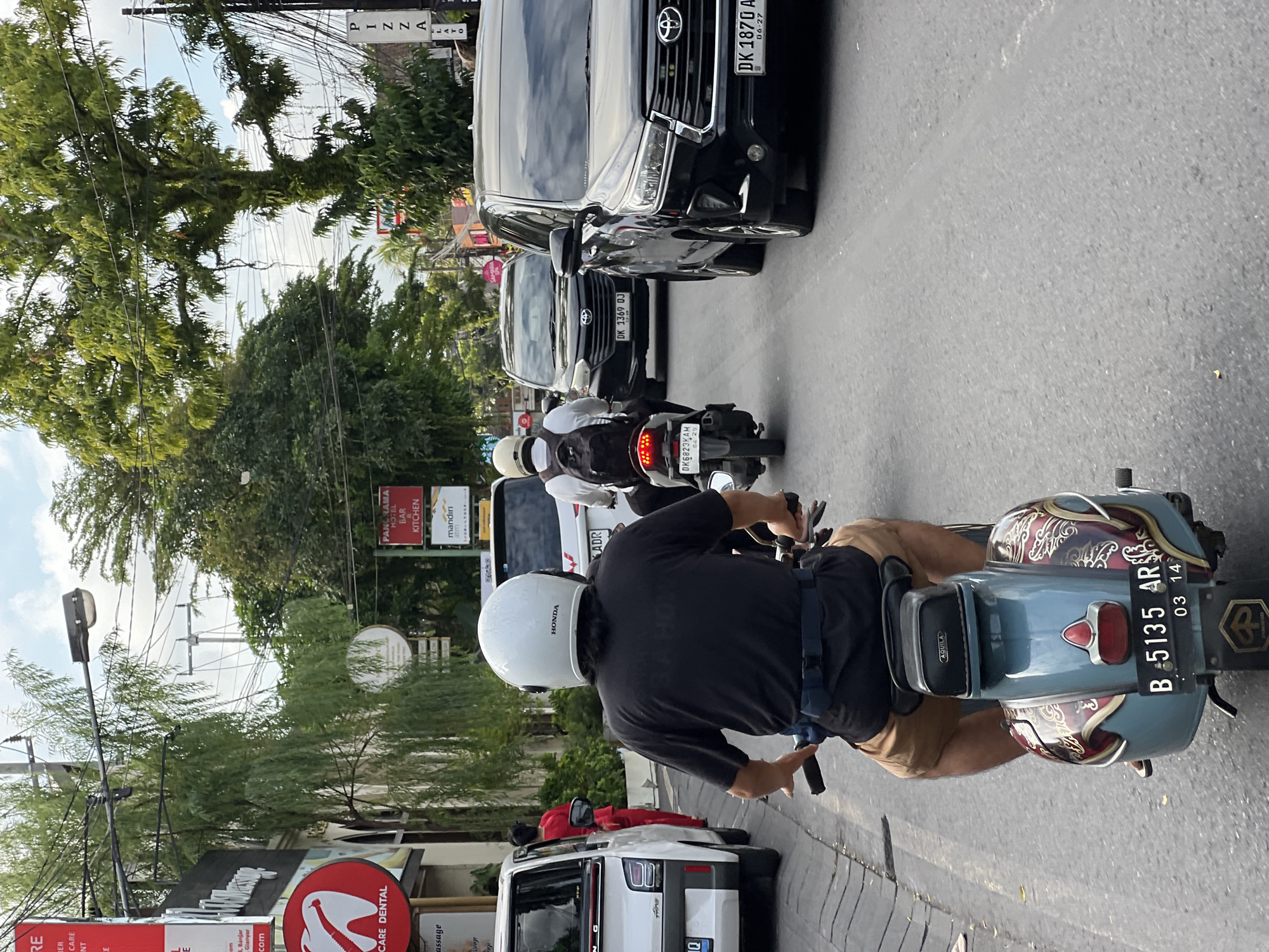 The image shows a street scene in Ubud District, Indonesia, with a focus on a person riding a blue Vespa scooter. The scooter has the license plate "B 5135 AR" and is moving along a road with other vehicles, including several cars (a black Toyota Innova with license plate "DK 1870 A", another black Toyota Innova with "DK 1369 OJ", and a white car). Other motorcycles can be seen ahead of the scooter.
The rider is wearing a black shirt, tan shorts, and a white helmet with the logo "HONDA". A black bag labeled "AQUILA" is attached to the rear of the scooter. The environment includes street signs, trees, power lines, and business signs, one of which reads "CARE DENTAL". A sign for "PANDRAMA HOTEL BAR KITCHEN" and "mandiri atm" can be seen. The road is paved and appears to be busy with traffic. Another license plate "DK6823KAM" is visible on one of the motorcycles in front. The presence of "PIZZALATO" is also noted.