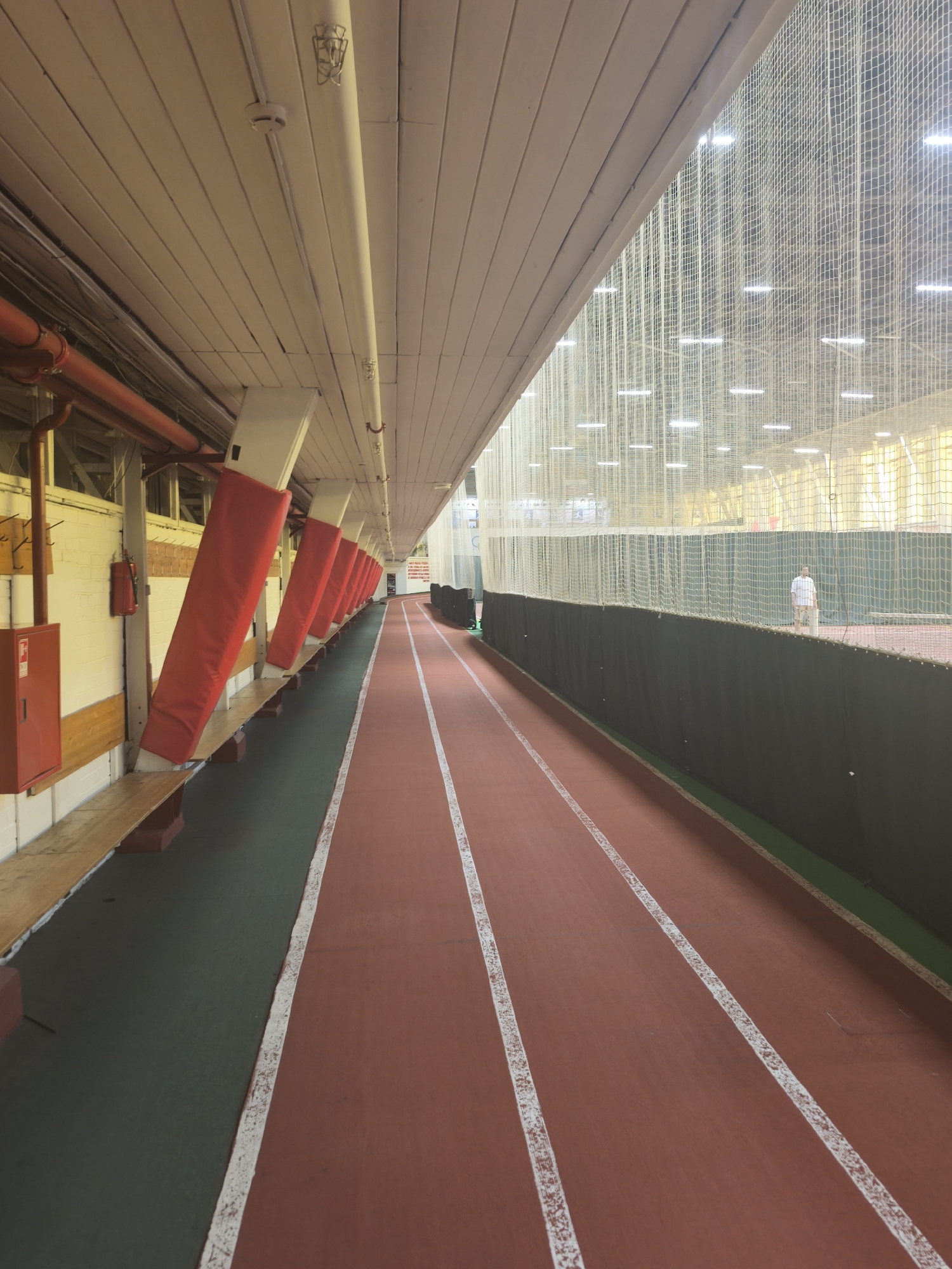 The image depicts an indoor running track within an athletic facility. The track is characterized by a reddish-brown surface divided into lanes by white lines. A green area borders the track on one side. On the left side of the image, there is a row of white support pillars covered with red padding. Behind the pillars, there is a wall with a bench running along it. A red fire extinguisher is mounted on the wall. Overhead, there is a wooden ceiling. On the right side of the image, a mesh net separates the track from an open area, where a person is standing in the distance. The lighting is artificial, provided by rows of overhead lights. The location is most likely in Saint Petersburg, Russia.