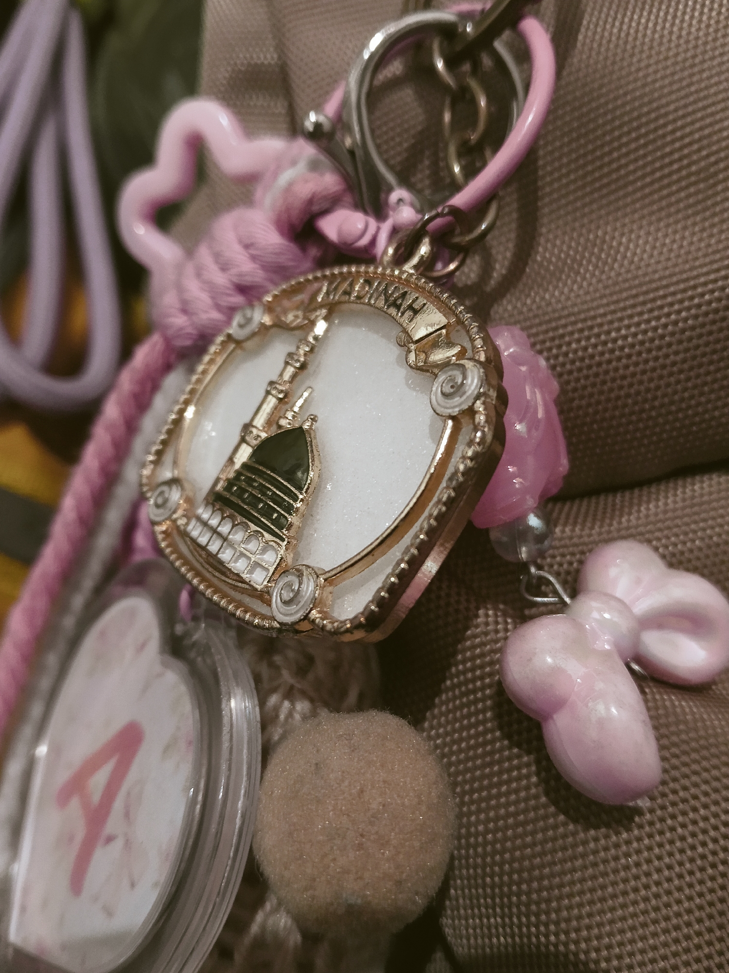 The image shows a close-up of a keychain with several decorative charms. The main charm is a circular, gold-colored pendant with an image of a building with a dome and minarets, likely a mosque, against a white, glittery background. The word "MADINAH" is inscribed in gold around the top edge of this pendant. Other charms include a pink heart-shaped plastic piece, a pink cord with a braided texture, a clear plastic heart-shaped charm with a pink letter "A" inside, a fuzzy brown ball, and a pink bow-shaped charm. These charms are attached to a silver-colored keychain ring and a pink chain. The background appears to be a textured brown fabric, possibly a bag. The lighting is dim, creating a soft focus effect. There are no people or specific landmarks visible that would indicate a location in Semarang, Indonesia.