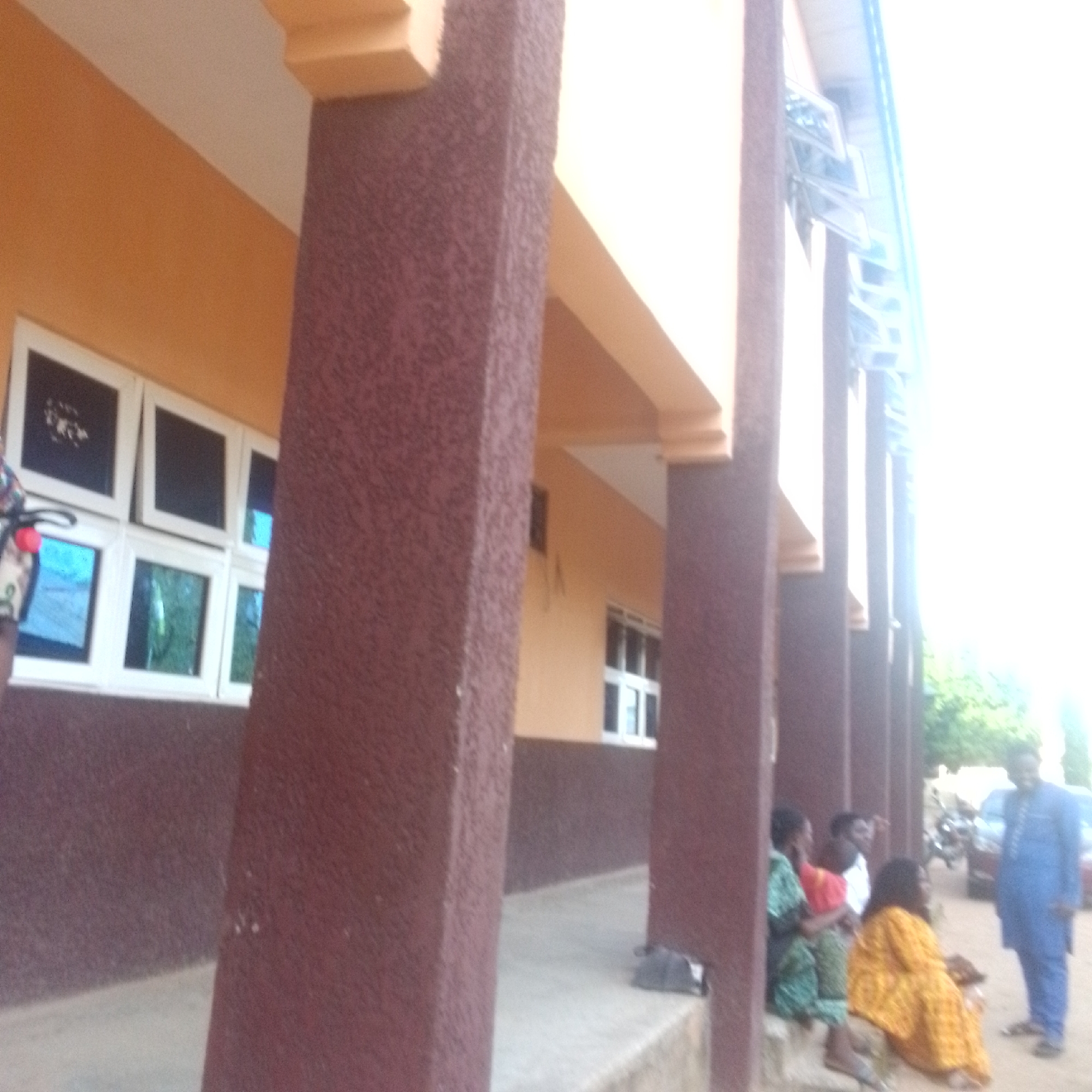The image shows an outdoor scene in Jalingo, Nigeria. A building with an orange facade and large, dark-paned windows dominates the left side of the frame. A rough-textured, reddish-brown column stands prominently in the foreground.
Several people are visible near the building's entrance. In the foreground, a group of individuals are seated on steps. One person wears a yellow and orange patterned garment. To their left, another individual is dressed in a green and white patterned outfit, and a third person is wearing a red top. Further to the right, a man in a blue traditional Nigerian outfit is standing.
The setting appears to be a public space, possibly a community center or a marketplace, given the building's architecture and the presence of people gathered. The lighting suggests it is daytime, with bright sunlight creating a washed-out effect on the right side of the image, hinting at a possibly sunny or hazy atmosphere. There are hints of greenery and trees in the background, along with what appears to be a motorcycle or scooter parked. No discernible text is visible in the image.