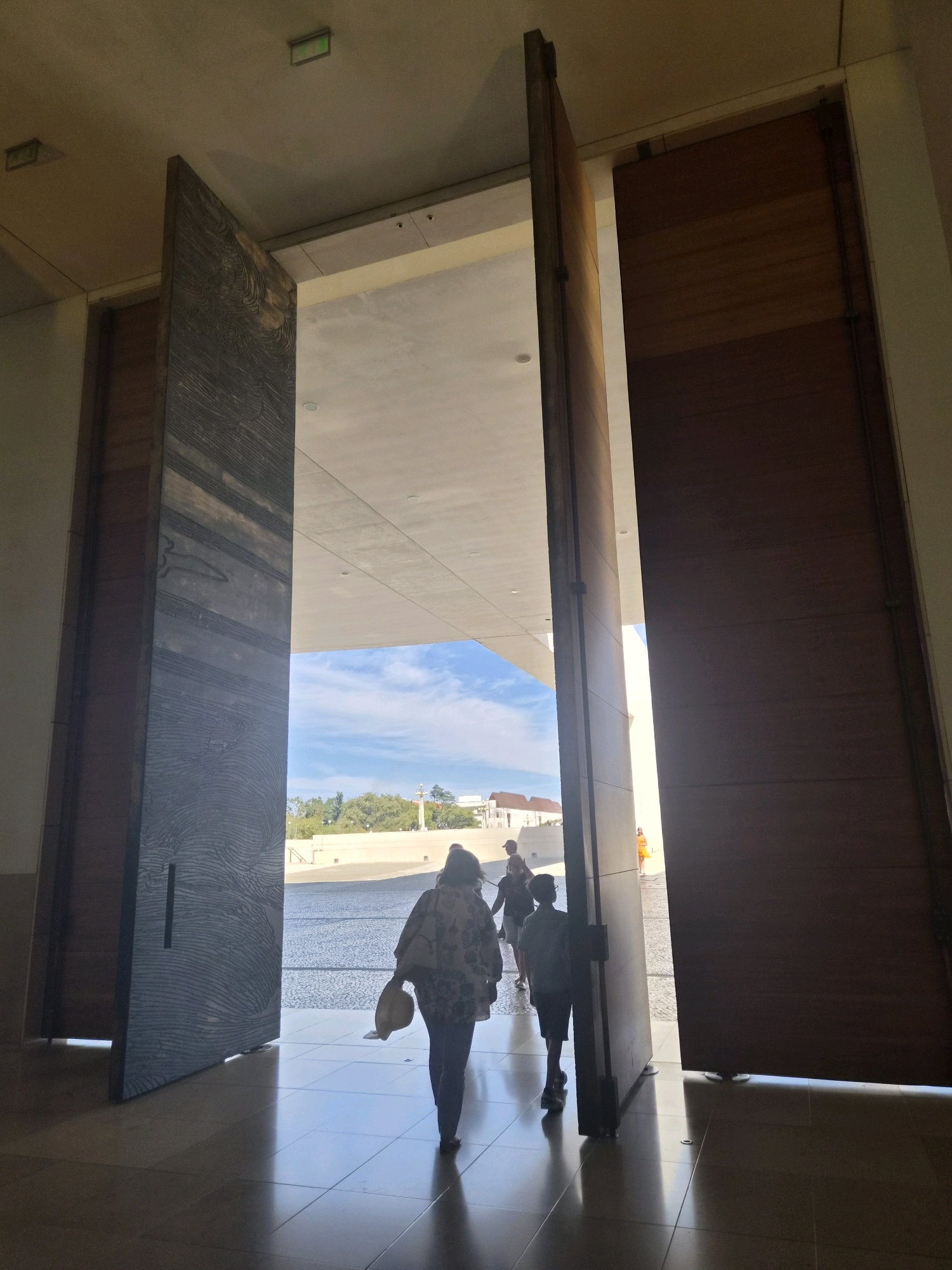 The image depicts a view looking out from an interior space through large, open wooden doors towards an outdoor plaza bathed in sunlight. Two large, imposing wooden doors are prominent in the foreground, one on the left with a textured, dark wood grain and the other on the right made of smooth, lighter wood. A woman and a boy are walking away from the camera, exiting the building through the opening between the doors, into the bright exterior. The woman, in the foreground on the left, wears a patterned jacket and carries a straw hat in her left hand. The boy, to her right, is silhouetted against the light. Other figures are visible further out in the plaza. The plaza itself appears paved and leads towards a body of water or reflective surface, with buildings and trees in the background under a blue sky with scattered clouds. The architectural style suggests a modern or contemporary building, likely part of a significant religious or public complex. Given the context of Fatima, Portugal, this scene is likely captured within the Sanctuary of Fátima, possibly near one of its entrances to the vast pilgrim square. The overall impression is one of transition from a shadowed interior to a sunlit, open space, with people moving towards it.