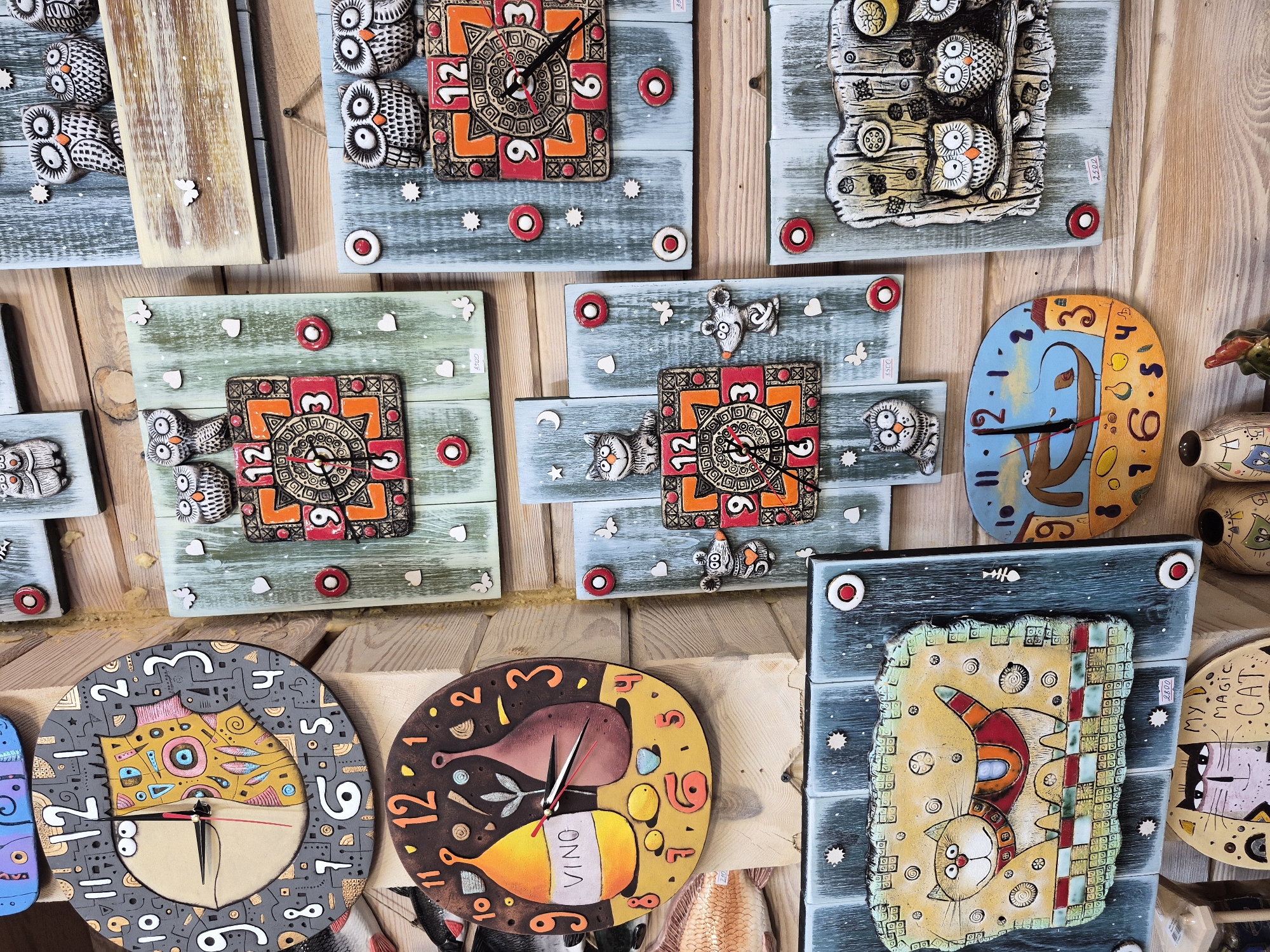 The image depicts a collection of handmade ceramic clocks displayed on a wooden wall, likely in a shop or market stall in Krasny Klyuch, Russia. The clocks are varied in design, featuring whimsical figures such as owls, cats, and mice, as well as more abstract patterns. Some clocks have a traditional round face with Arabic numerals, while others have square or irregularly shaped designs.
Prominent among the clocks are several with owl motifs. One large round clock, in the upper left, is gray with black numbers and a colorful, abstract face. Below it, a circular clock with a brown background features two wine bottles and the word "VINO". To the right, a series of square or rectangular clocks are arranged vertically. These include clocks with owls, hearts, and decorative patterns in oranges and reds. One clock features a charming cat in a colorful scarf, and another shows a blue background with a boat and a bird. A clock with a mouse figure is also visible.
The wooden wall serves as a rustic backdrop, with visible grain and texture. The lighting appears to be natural or from overhead, casting a soft illumination on the objects. There are no people visible in the image. The scene is static, presenting a still life of decorative items. Small price tags are visible on some of the clocks.