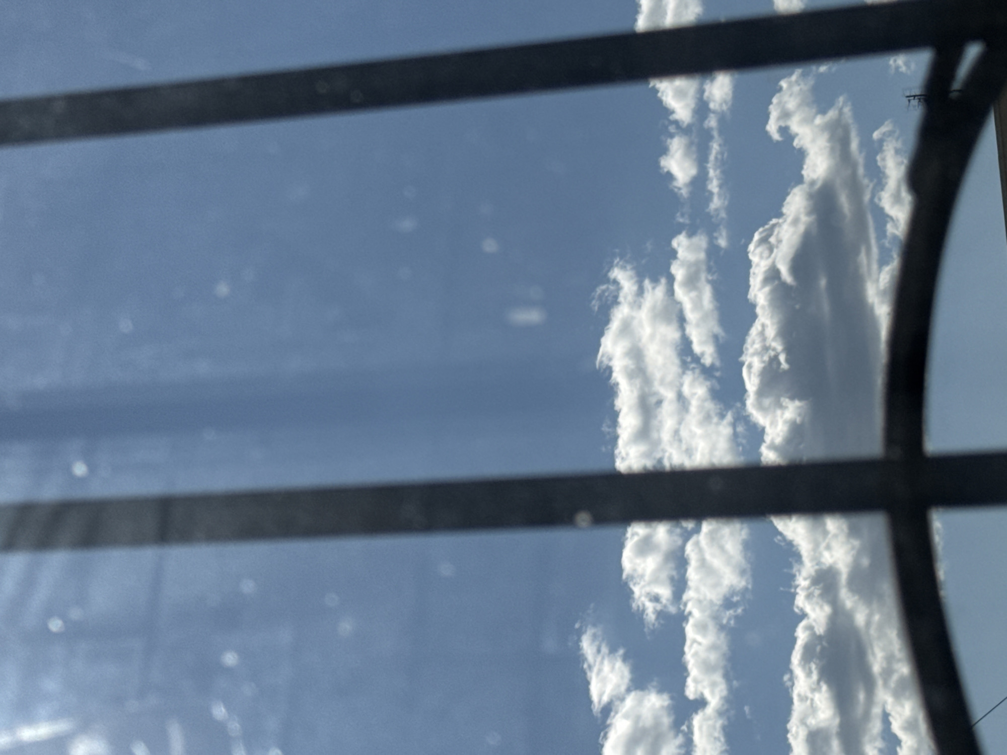 The image shows a view of the sky through what appears to be a barred window or fence. The sky is predominantly blue with scattered white cumulus clouds. The bars are dark and vertical, with a curved, dark object in the foreground. The image is taken from an indoor perspective looking outwards. No people or specific structures within the city of Zaokskiy, Russia are visible.