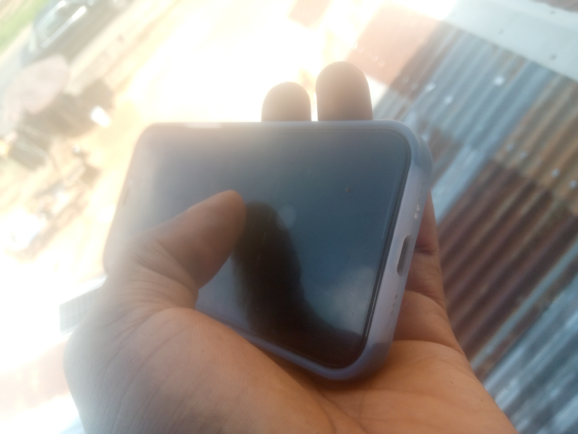 A medium-dark skinned right hand holds a smartphone encased in a light blue or lavender silicone-like case. The phone's screen is dark, possibly off, with a faint, circular light reflection near its center. Visible features of the phone case include a cutout on its right side, likely for buttons, and a series of small embossed dots. The background is brightly overexposed on the left, with indistinct, blurred shapes suggesting distant structures or vehicles under intense sunlight. To the right, a corrugated metal roof extends diagonally, displaying its characteristic parallel ridges and reflective surfaces, indicating an outdoor setting in Jalingo, Nigeria.