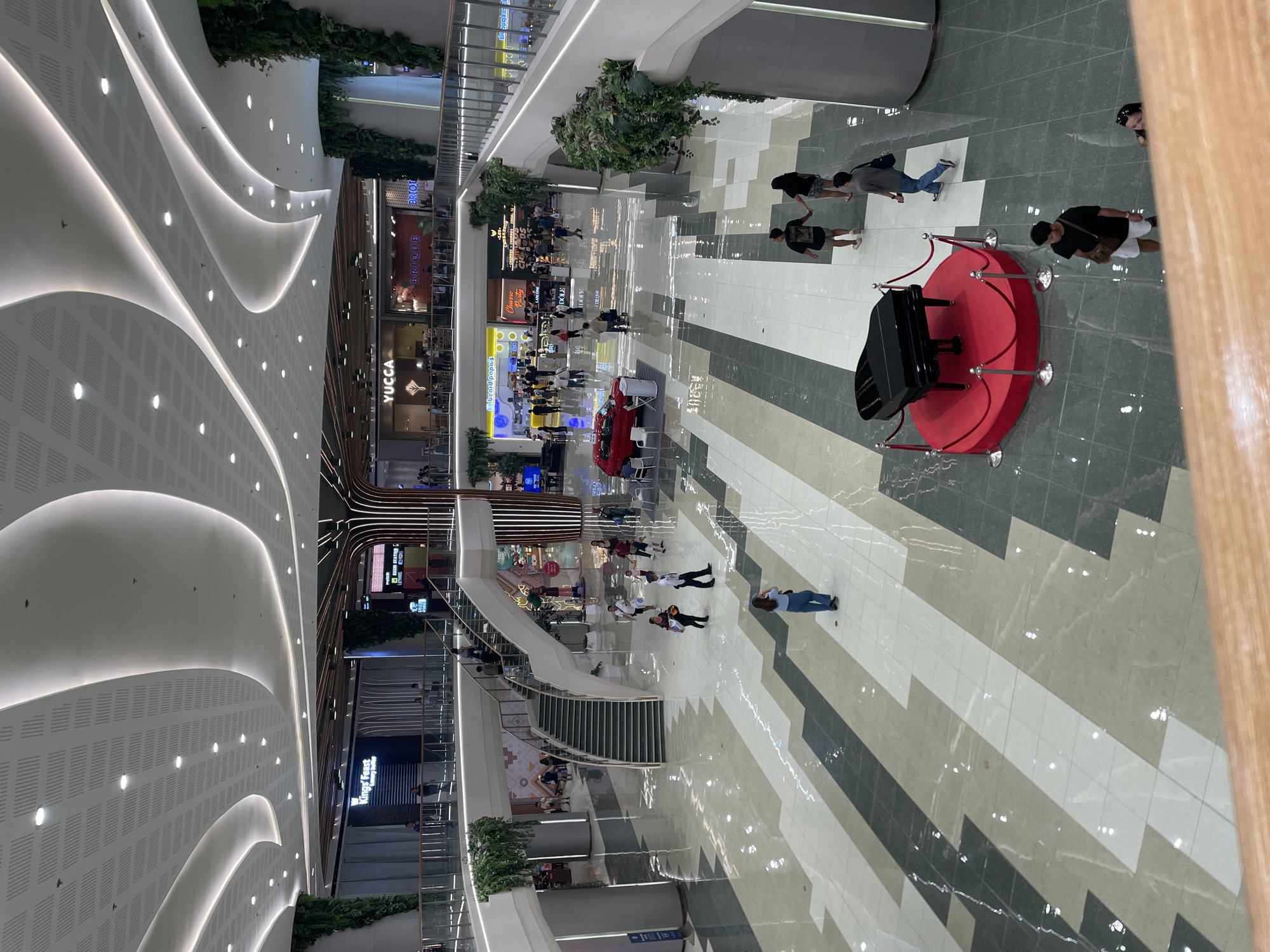 This image captures a wide, high-angle view of the interior of a modern, multi-level shopping mall atrium in Davao City, Philippines.
The mall features a high, white, curvilinear ceiling with integrated strip lighting and numerous recessed circular lights, interspersed with sections of perforated acoustic panels in a square grid pattern. Several large structural pillars and portions of the balustrades on upper levels are adorned with extensive green vertical gardens or hanging foliage.
The ground floor, which is highly polished and reflective, showcases a large geometric tile pattern in alternating shades of white, light grey, dark grey, and light green/beige. In the lower foreground, a black grand piano is prominently displayed on a circular red platform, enclosed by red velvet ropes and silver stanchions. Further into the ground floor, a red compact car is exhibited on a white platform, accompanied by two white chairs and a small table.
A wide, double-flight staircase with dark treads and white risers, featuring a central landing, connects the ground floor to the first upper level. Glass balustrades with metal top rails line the edges of the upper floors. A large, multi-story, stylized decorative tree structure with integrated vertical lighting stands centrally, rising through multiple levels.
Numerous individuals are observed navigating the mall, walking, standing, and congregating. They are dressed in casual attire. Visible storefronts on the upper levels include "King's Feast Luxury Buffet," "YUCCA," "BRIQUE," "beard papa's," "Churro Party," "Lancôme," and "SUSHI STATION G." The overall environment is well-lit and appears spacious.