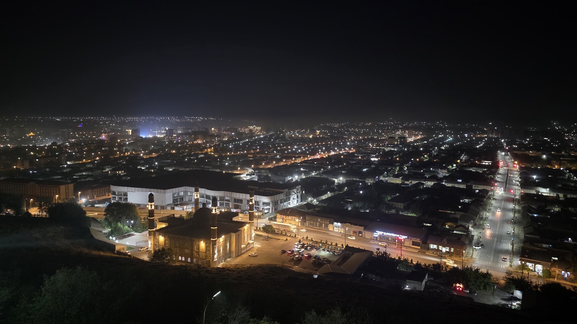 This is a high-angle night-time photograph of a city, likely Osh City, Kyrgyzstan, featuring a mosque and a large building complex in the foreground, with a sprawling urban landscape extending into the distance. The mosque, illuminated by warm lights, has two minarets and a central dome structure. Adjacent to it is a modern, multi-story building with a white facade and dark windows, possibly a shopping center or administrative building.
The foreground also shows a slope covered in vegetation and a street with parked cars. The urban area beyond is a dense network of buildings, illuminated by streetlights, traffic lights, and internal building lights, creating a blanket of light across the landscape. A main road with moving vehicles is visible, with traffic signals casting red and green glows. The distant skyline is dotted with more lights, suggesting a large and active city. The sky is dark, with a slight haze, common in urban environments at night. There are no visible people in the image, only evidence of human activity through the illuminated structures and vehicles.