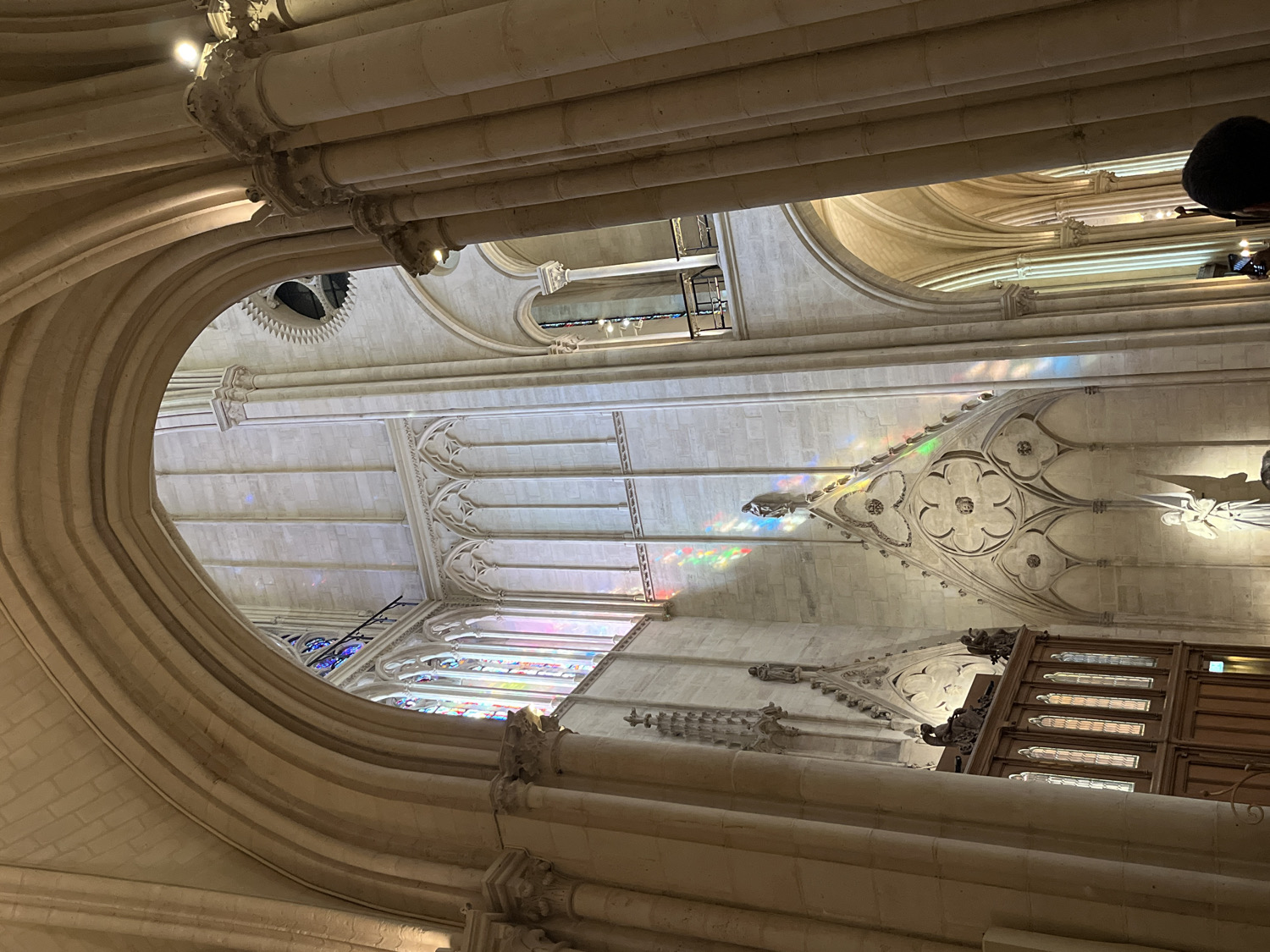 This image captures the majestic interior of a large, ornate stone building, likely a Gothic or Neo-Gothic cathedral or church in Paris, France. The perspective looks upwards, emphasizing the immense height and intricate details of the architecture.
Dominating the frame are the light-colored, cream-hued stone arches and ribbed vaults that form the ceiling structure. Thick, multi-layered arches in the foreground frame the view, revealing the complex construction and decorative carvings, including capitals and possibly gargoyles or small statues integrated into the stonework. High walls feature further decorative elements, such as trefoil and quatrefoil patterns.
A significant feature is the play of natural light. Sunlight streams through unseen stained glass windows, casting vibrant, rainbow-colored reflections onto the stone walls and decorative structures, creating a breathtaking visual effect. These colorful patches are prominent on the mid-ground architectural elements. Through various arches, glimpses of additional architectural details, including more stained glass windows and high-arched ceilings, are visible further into the building.
In the upper right corner, the top of a person's head and shoulders, with dark hair or clothing, is partially visible, indicating an observer looking up at the grand interior. On the lower right, a dark wooden structure, possibly an organ console or choir stalls, with rectangular panels and windows, is partially cropped. Small light fixtures are also mounted on some of the arches and walls.
The scene conveys a sense of grandeur and awe, suggesting a daytime visit to appreciate the architectural splendor and the beautiful interplay of light and stone. No visible text is present.