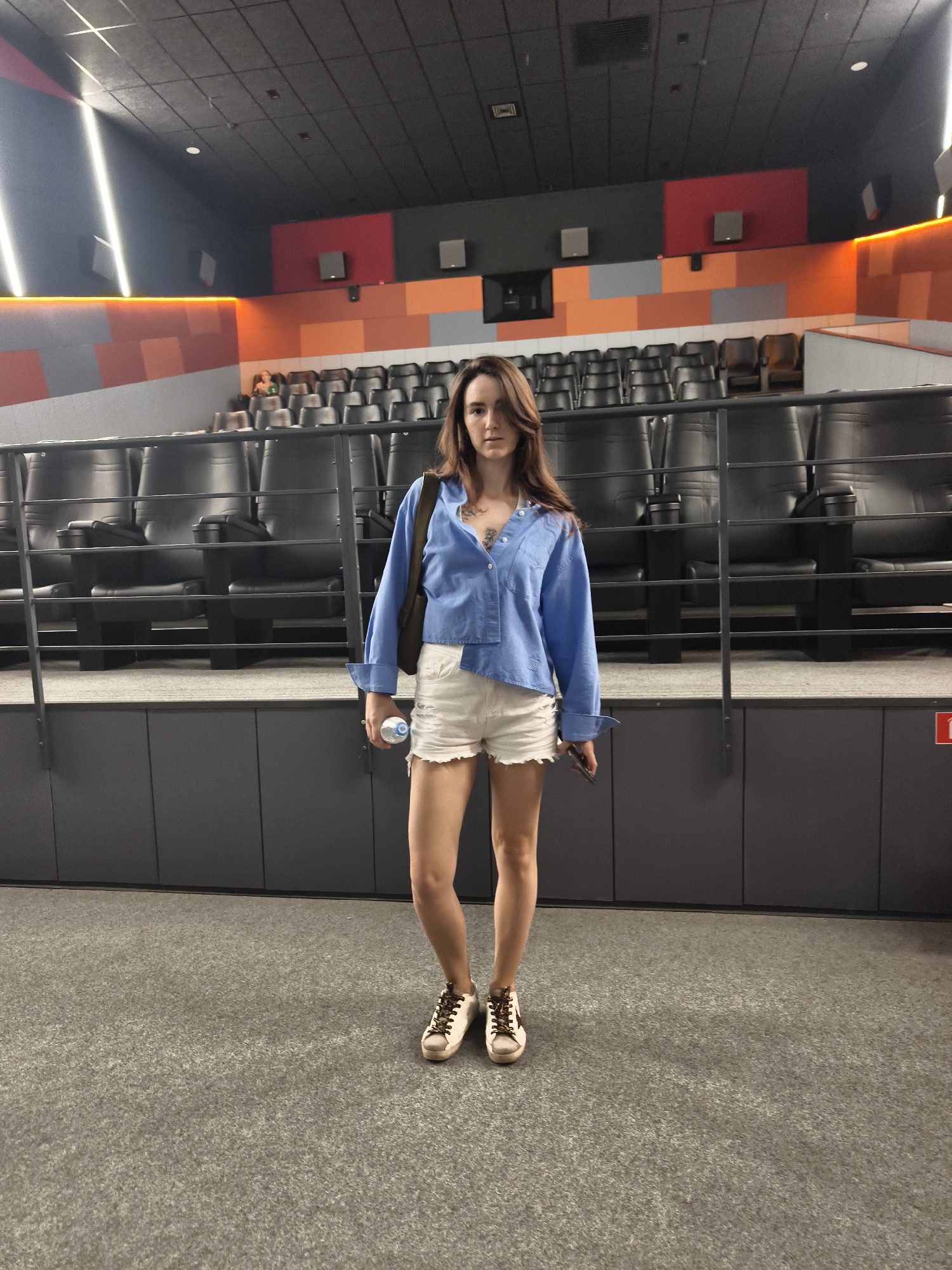 A young adult female stands facing the camera in an empty movie theater. She is wearing a light blue unbuttoned shirt over a dark camisole, white distressed denim shorts, and white sneakers with brown laces and a gold star detail. She holds a clear plastic water bottle in her right hand and her left hand is slightly out of frame. She has long brown hair that falls over her shoulders and a tattoo is visible on her chest. The theater seating is visible behind her, with dark grey chairs arranged in rows. The walls of the theater are decorated with geometric patterns in orange, grey, and red. The lighting in the theater is dim, with bright white linear lights on the ceiling. The floor is covered in grey carpet. The image does not contain any visual cues to indicate it was taken in the city of Tyumen, Russia.