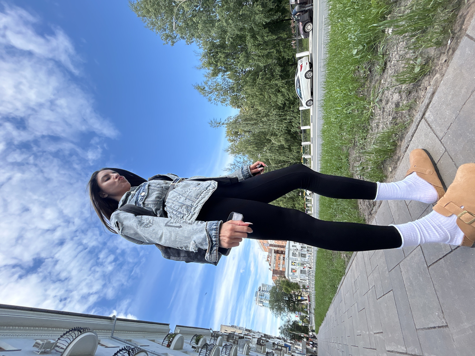 A low-angle, full-body shot captures a young woman standing on a paved sidewalk. She is wearing a light-wash denim jacket with a graphic print, black leggings, white crew socks, and tan slip-on shoes. Her dark hair is down, and she is holding a smartphone in her right hand. She appears to be making a kissy face, looking up towards the camera.
The background reveals a vibrant scene. A bright blue sky with scattered white clouds dominates the upper portion of the image. Below the sky, a lush green tree line runs horizontally, separating the sky from a city street. Cars are visible on the street, including a white sedan and a darker vehicle. A white fence separates the grassy area from the street. In the distance, urban buildings with multiple stories can be seen.
The paving stones of the sidewalk are laid in a repeating pattern. The overall lighting suggests it is daytime, likely a sunny afternoon, given the clear sky and shadows. The setting appears to be an urban environment in Novokuybyshevsk, Russia. The woman's relaxed posture and facial expression suggest a casual, possibly playful moment. No visible text is present in the image.