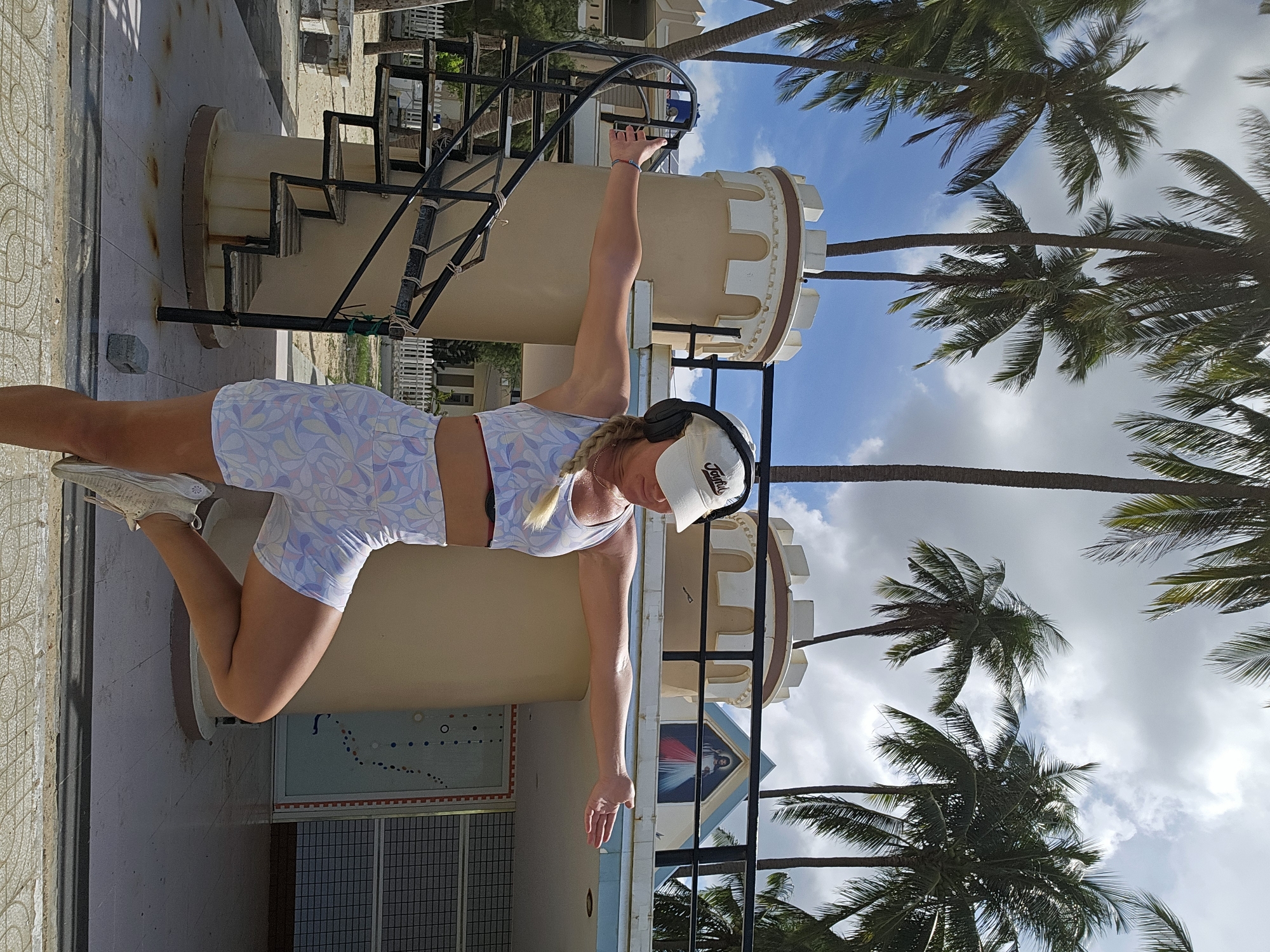 A woman stands on one leg with her arms extended horizontally in a yoga pose. She is wearing a white baseball cap, headphones, a sports bra, and shorts with a floral pattern. The woman is positioned in front of a cream-colored tower with a white crenellated top, resembling a castle. To the left of the tower is a black spiral staircase. Behind the tower, there is a building with a blue roof and a triangular section adorned with a painting of Jesus Christ. Palm trees are visible in the background, suggesting a tropical or subtropical location. The ground in the foreground is paved with tiles and concrete. The sky is blue with scattered clouds. The scene appears to be outdoors, likely in a public area with a unique architectural style and natural elements. The image is taken from a low angle, emphasizing the height of the tower and the palm trees. The overall setting is consistent with the appearance of the Mũi Né coastline in Vietnam, known for its resort developments and beachside attractions.
