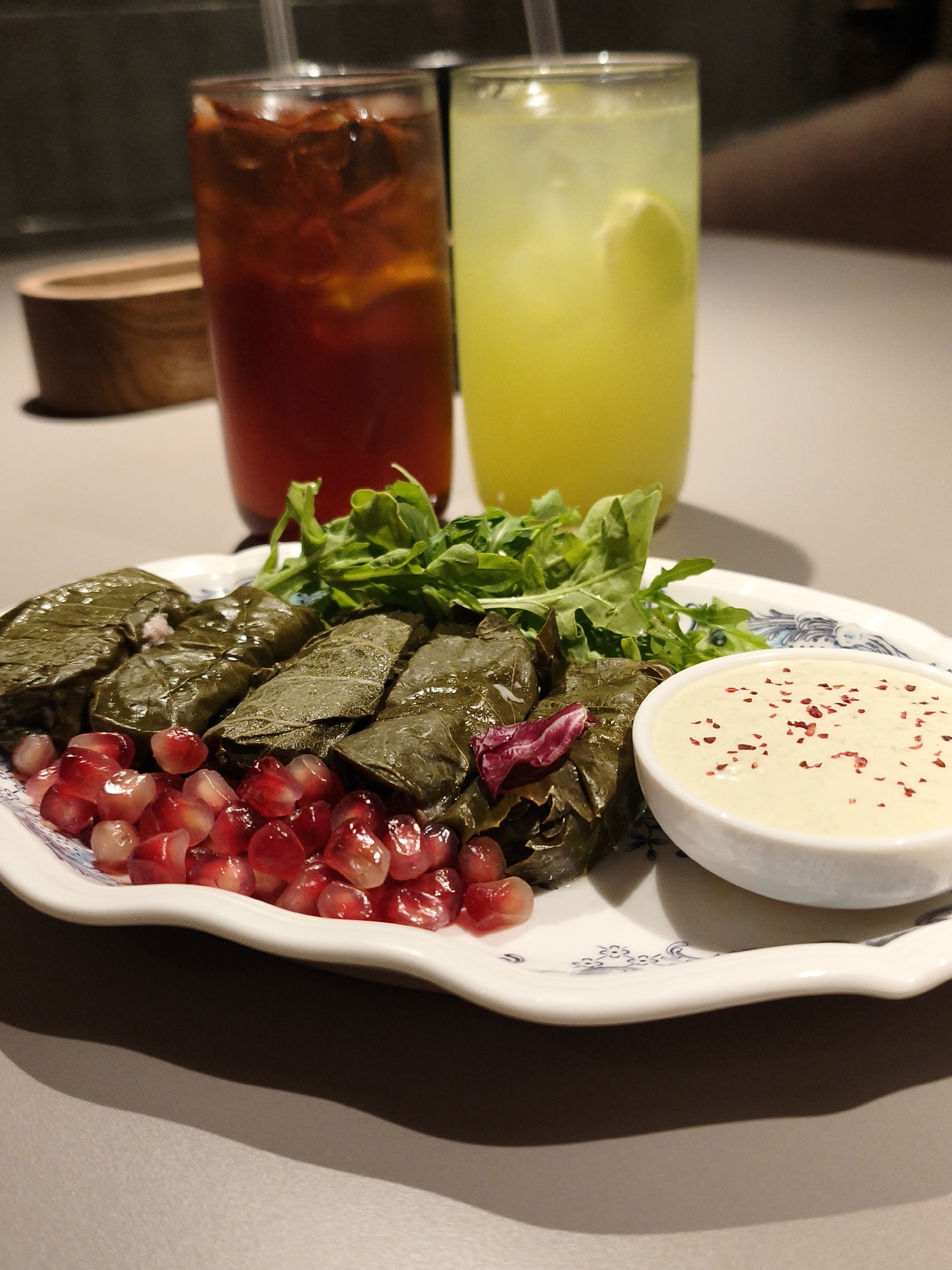 A close-up, eye-level shot captures a plate of dolmades served with a side of creamy dip and pomegranate seeds, accompanied by two tall glasses of drinks. The dolmades, wrapped in dark green grape leaves, are arranged neatly on a white plate with a blue decorative rim. Scattered across the plate are vibrant red pomegranate seeds, adding a splash of color. A small white bowl holds a light-colored, creamy dip, sprinkled with red flakes, likely sumac or chili flakes. A cluster of fresh green arugula sits atop the dolmades.
In the background, two tall glasses are visible. The glass on the left contains a dark red liquid, possibly iced tea or a fruity beverage, with ice and possibly mint. The glass on the right holds a pale yellow liquid, likely lemonade or a citrus-based drink, with a slice of lemon floating within. Both drinks have straws.
The setting appears to be indoors, likely a restaurant or home dining area, with a neutral-toned tabletop. The lighting suggests it could be daytime, possibly late afternoon, given the warm, soft light. The focus is on the food, with the drinks and background slightly out of focus, creating a shallow depth of field. The overall mood is inviting and suggests a pleasant dining experience. No visible text is present in the image.