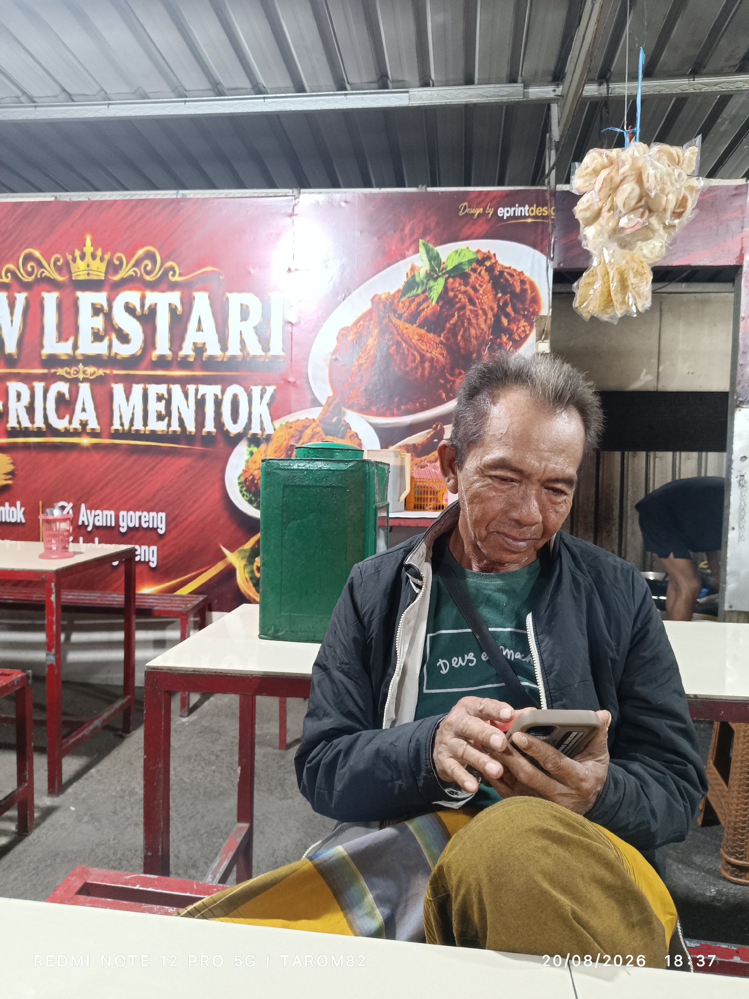 A man sits at a table in an outdoor food stall, looking at his phone. He is wearing a dark jacket over a green t-shirt and a yellow and brown patterned sarong. In the background, a large banner displays "W Lestari Rica Mentok" and images of food. There are red tables and chairs, and a green water dispenser. The scene appears to be in Bringinbendo, Indonesia. The time stamp indicates the photo was taken on 20/08/2026 at 18:37.