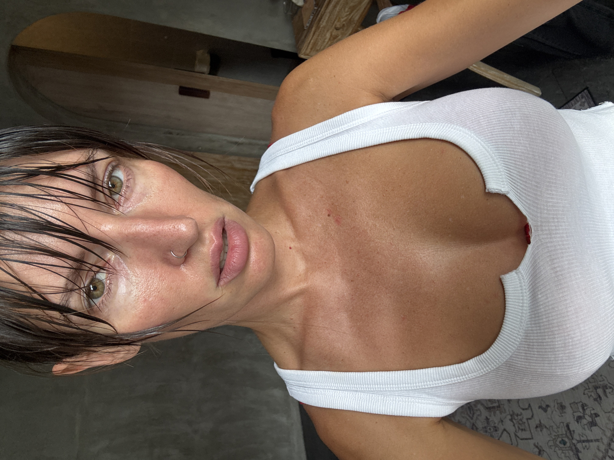 The image shows a close-up, low-angle shot of a woman. She has wet, dark hair that is partially obscuring her face. Her eyes are open and looking directly at the camera, with a neutral expression. She is wearing a white ribbed tank top that is low-cut, revealing her décolletage and the upper part of her breasts. A thin silver hoop is visible in her nostril. Her skin appears slightly sweaty or dewy.
The background is blurred and includes muted tones of wood and dark grey. There are hints of furniture and possibly a rug on the floor. The lighting suggests an indoor setting, possibly with natural light coming from an unseen source. The overall mood is intimate and direct due to the close-up framing and the woman's gaze. There is no visible text in the image. The scene is set in North Kuta, Indonesia.