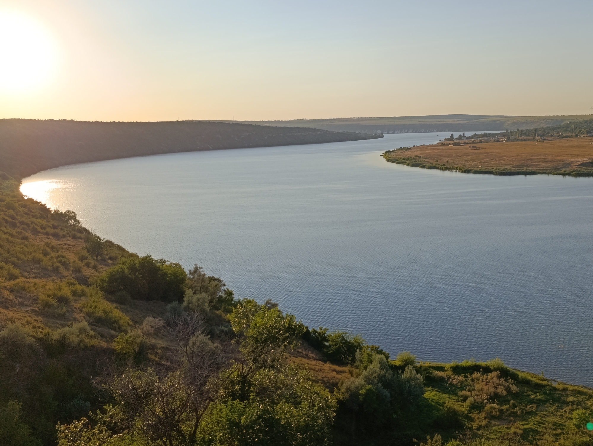 The image depicts a wide river bend with a sunlit bank on the left and a more populated bank on the right. The sun is setting on the left, casting a warm glow across the water and the lush, green hillside dotted with trees and bushes. The right bank features a dry, golden field with scattered buildings and trees, suggesting a rural or semi-rural settlement. A small village or cluster of buildings is visible, and a wind turbine stands on the horizon. The river itself is calm with subtle ripples, reflecting the pale blue of the sky. The overall scene is serene and natural, characteristic of a scenic landscape near Roghi, Moldova, likely in the evening.