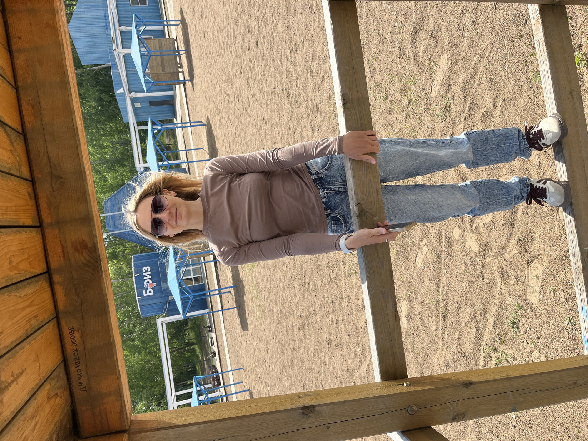 This image depicts a young woman standing outdoors on a sunny day, framed by a wooden structure, possibly a gazebo or pergola. She has blonde hair and is wearing sunglasses, a long-sleeved light brown top, blue jeans, and white/grey sneakers with dark laces. A ring is visible on her right hand, and a white-strapped watch is on her left wrist. She is looking directly at the camera with a neutral expression, holding onto a vertical wooden beam.
The setting appears to be a recreational area, possibly a beach or park, characterized by a sandy or dirt ground with sparse green vegetation. In the background, several blue, shed-like buildings with white trim are visible. One of these buildings prominently features the Cyrillic text "Бриз" (Briz), meaning "Breeze." In front of these buildings, there are blue metal frames, which could be for sunshades or benches. Green trees and foliage can be seen in the distance, suggesting a natural environment.
A notable detail is the text carved or written on a horizontal wooden beam in the lower left foreground: "ДМ ЧАЙКОВСКИЙ 02.000032". The word "ЧАЙКОВСКИЙ" (Chaikovsky) matches the given location context of Chaikovsky, Russia. The scene conveys a bright, clear day, likely late morning or afternoon, with strong sunlight casting shadows.