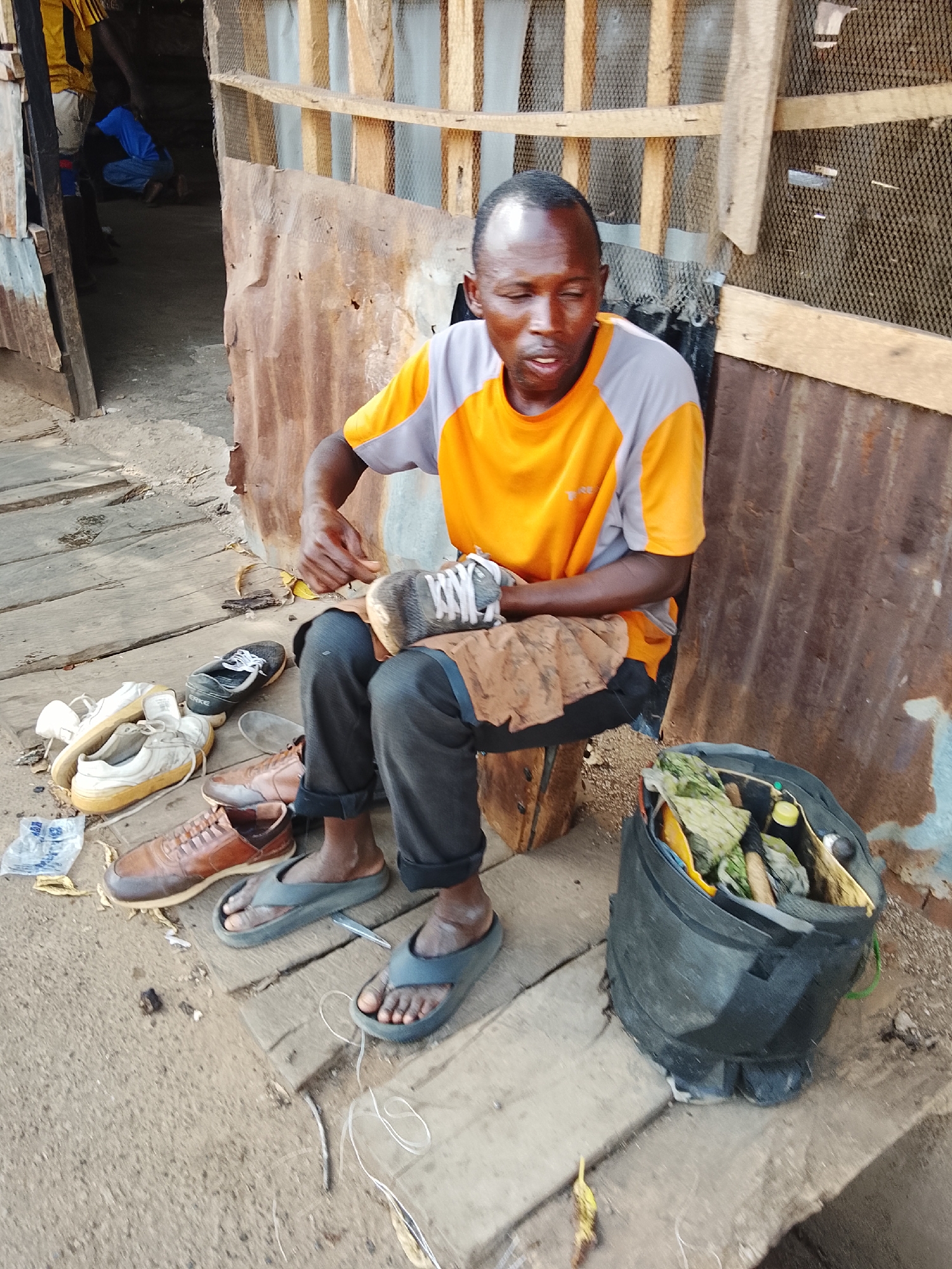 A medium shot captures a shoemaker, an adult male, seated and engaged in his craft in Akwanga, Nigeria. He is positioned on a low wooden stool or box on a platform of wooden planks, surrounded by footwear and his tools. The man wears an orange and grey short-sleeved t-shirt, dark trousers, and dark flip-flops, with a light brown cloth draped over his lap. His gaze is directed downwards, focused on a grey sneaker he holds in his hands, suggesting he is actively mending or preparing it.
Numerous pairs of shoes are laid out on the wooden planks beside him, including light-colored sneakers, brown leather lace-up shoes, and brown slip-ons. One black sneaker clearly displays the brand "ERKE." To his right, a black bag, resembling a backpack, stands open, revealing various tools, bottles, and materials pertinent to shoe repair. A length of white string or wire lies on the ground near his feet.
The setting appears to be an informal outdoor or semi-outdoor workshop or stall. Behind the shoemaker, a wall constructed from rusty, corrugated metal sheets rises, topped with a wooden frame and mesh or netting. To the left, a dark doorway leads into a less discernible interior, where glimpses of other individuals, including one in a blue shirt, can be seen. The ground is a mix of dirt and sand. The scene is illuminated by natural daylight, indicating it is daytime. The man's expression is one of concentration. A discarded white plastic bag with some indistinguishable blue text is also visible near the shoes.