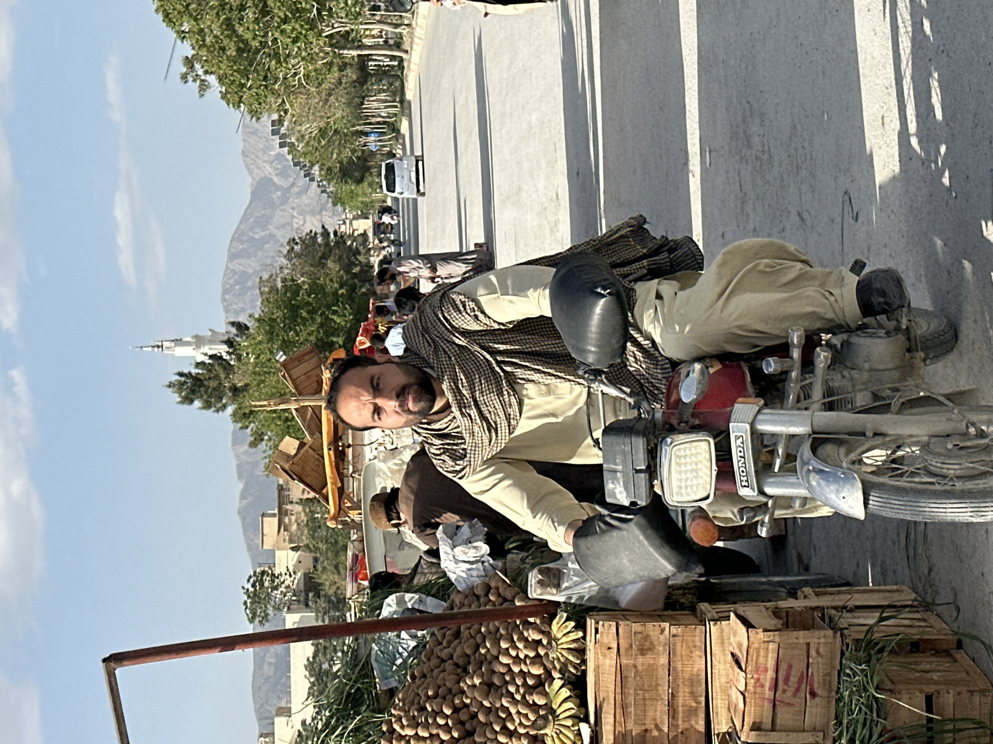 A man sits on a red Honda motorcycle in Quetta City Tehsil, Pakistan. He wears a tan outfit and a patterned scarf. Crates of potatoes, bananas, and greenery sit in front of the motorcycle. In the background, there is a road, trees, buildings, and mountains under a blue sky with a few clouds. A tall white building stands out.