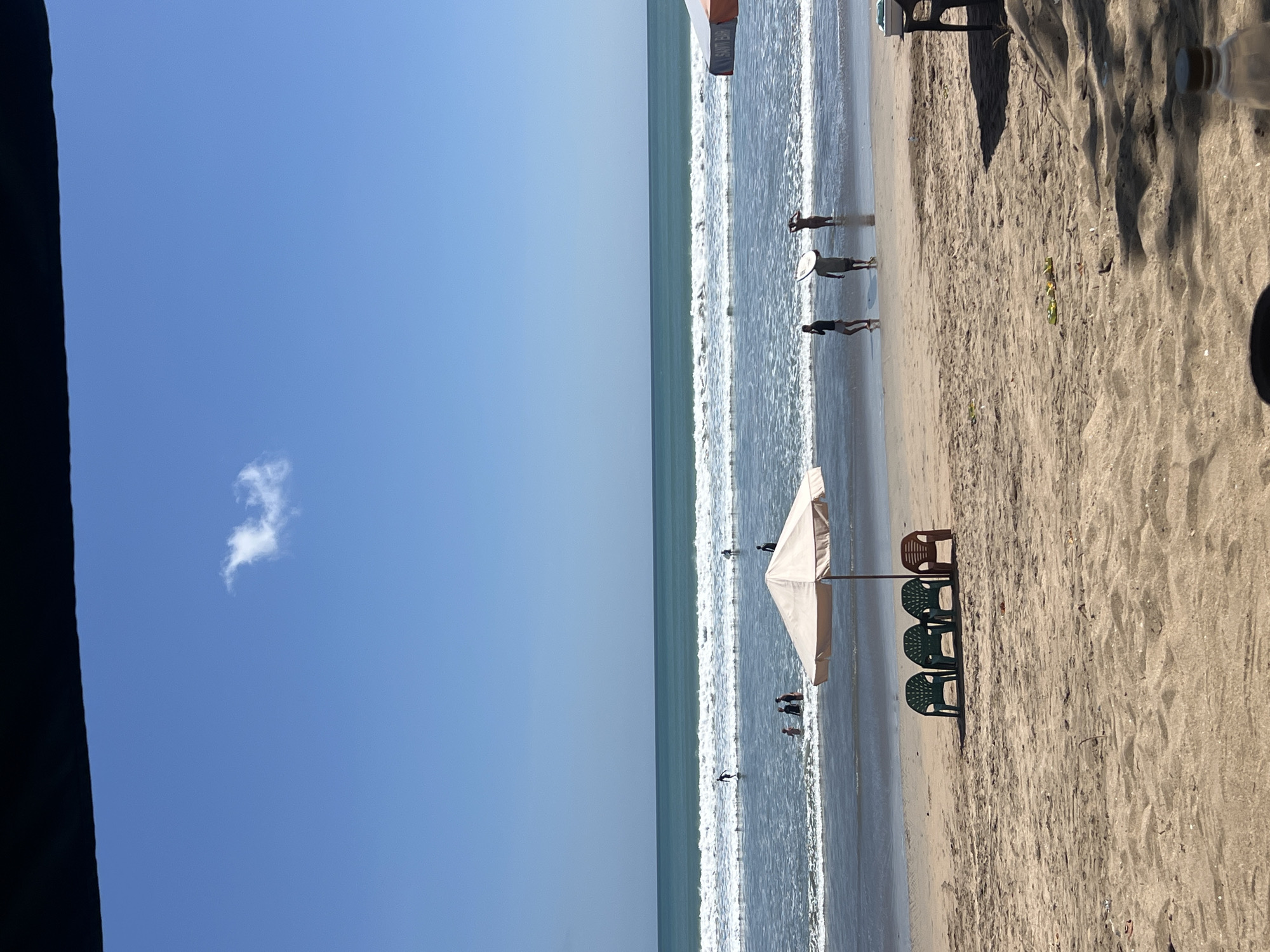 The image captures a sunny day at Kuta Beach, Indonesia, with clear blue skies and a single wispy cloud. The ocean is calm with gentle waves breaking on the shore. Several people are visible in the water, some appearing to be surfing or wading. On the sandy beach, a beige beach umbrella shades four plastic chairs, three green and one brown. Further down the beach, other beach umbrellas and a table with chairs are partially visible, suggesting a lively beachside setting. The sand is light brown with some footprints and scattered debris. The scene conveys a relaxed atmosphere typical of a popular tourist destination.
