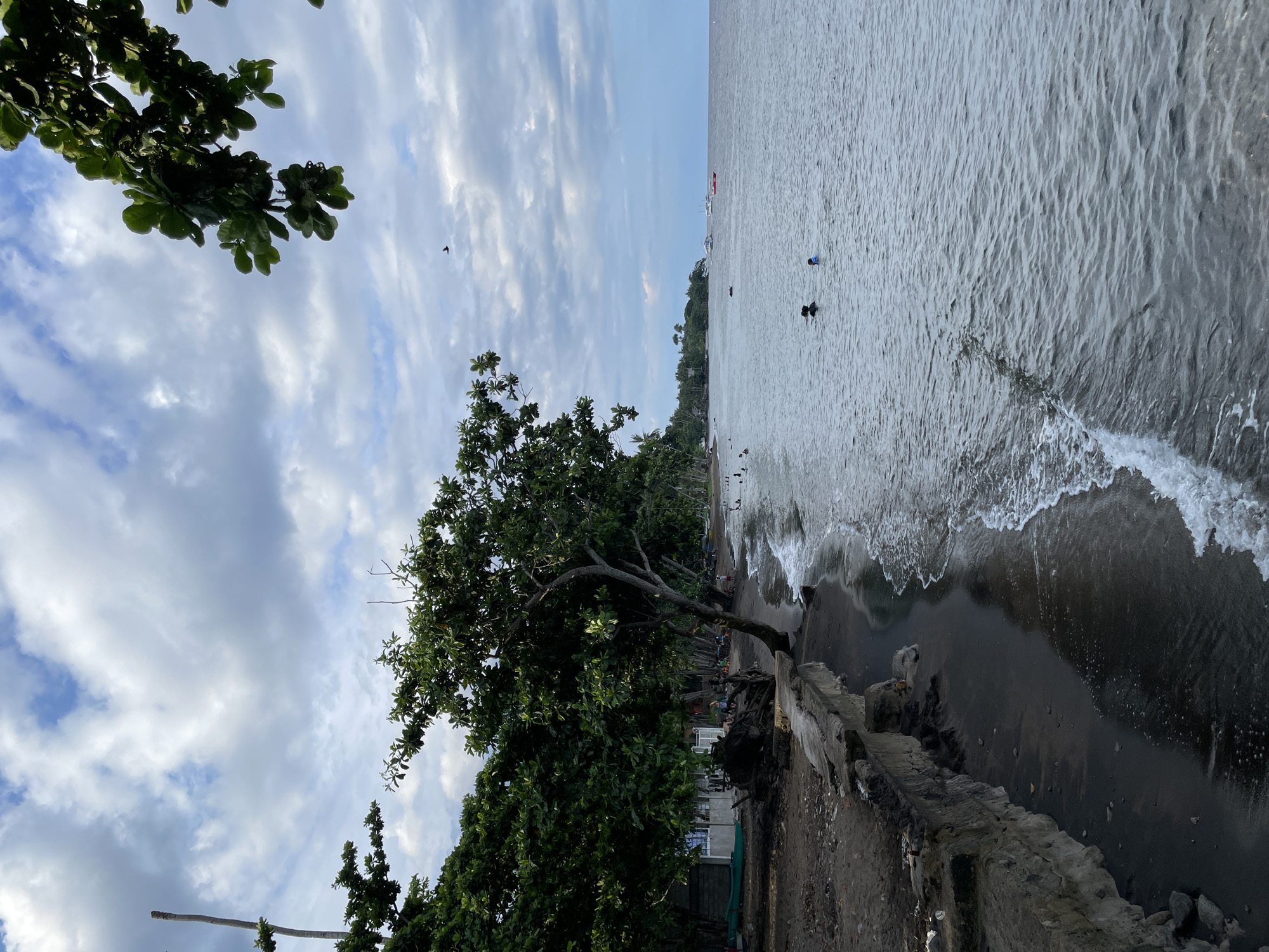 The image depicts a tropical beach scene in Dauin, Philippines. The foreground features dark volcanic sand with waves gently lapping at the shore. A concrete embankment, showing signs of wear and tear, lines the edge of the beach in the left foreground. Lush green trees with thick foliage overhang the embankment and beach, providing shade.
Further along the beach, several people are visible. Some are walking or sitting near structures that appear to be small buildings or huts. A green boat is beached on the sand near one of these structures. In the water, a few individuals are swimming or wading, their heads and shoulders visible above the surface.
The sea extends to the horizon, meeting a sky filled with a mix of white and grey clouds, suggesting it might be late afternoon or early evening. Several boats, including what appear to be outrigger boats and a red object that could be a buoy or small vessel, are present on the water in the distance. A lone bird is captured in flight against the cloudy sky. The overall impression is of a relaxed, coastal environment with evidence of local life and activities.