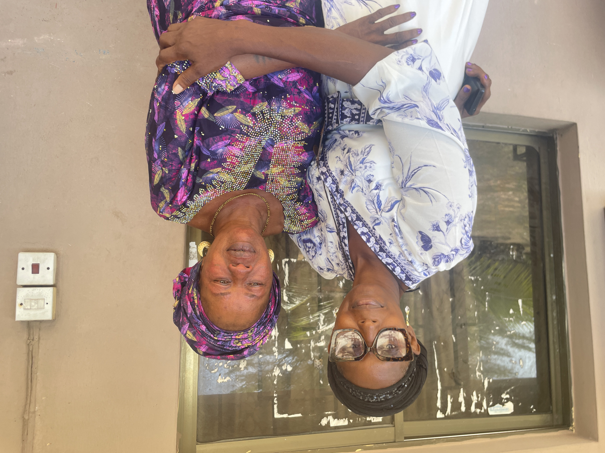 Two women are seated, posing for a photograph. One woman, on the left, is wearing a purple and pink patterned dress, while the other is wearing a white and blue patterned dress. Both are wearing head wraps. The woman on the right is wearing glasses. They are sitting indoors near a window. The location is likely Badagry, Nigeria, based on the prompt. A light switch is visible on the wall beside them.