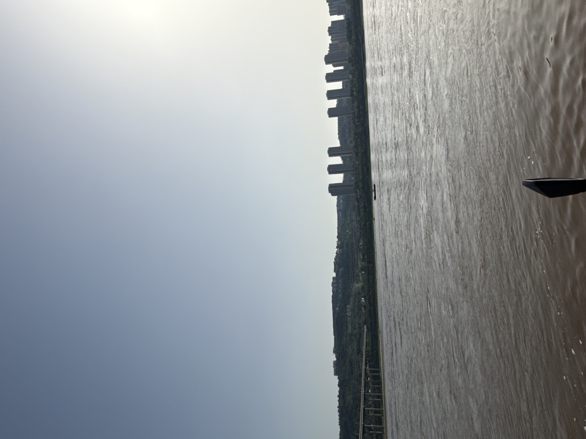The image captures a wide, expansive view of a riverine landscape in Jiangjin, China, presented from a perspective that includes the sky, a distant cityscape along the riverbank, and the river itself.
The upper portion of the frame is dominated by a bright, somewhat hazy sky, transitioning from a luminous white at the top to a soft, light blue-grey further down, suggesting either a very bright or slightly overcast daytime. The sun appears high, creating diffused light across the scene.
Along the horizon, separating the sky and the water, lies a strip of land. This land is characterized by green, vegetated hills, upon which numerous tall, modern residential buildings are constructed, forming a dense urban skyline. These structures appear somewhat indistinct due to distance and atmospheric haze. On one of the green hillsides, faint white characters, likely Chinese, are visible, possibly indicating a landmark or name. A long, dark structure, potentially a pier or an embankment, is visible along the river's edge on the left side of the frame.
The lower portion of the image is occupied by a large body of water, consistent with a wide river. The water is a murky, brown-grey color, characteristic of sediment-laden rivers. Its surface shows numerous ripples and small waves, indicating movement. In the bottom right corner, a dark, angular object, possibly part of a boat, ferry, or platform, extends into the frame from the water.
The scene depicts a blend of natural landscape and urban development typical of riverside cities. The bright, hazy conditions suggest a daytime setting, possibly late morning or early afternoon, with no strong shadows evident. No specific activity or individuals are clearly visible.