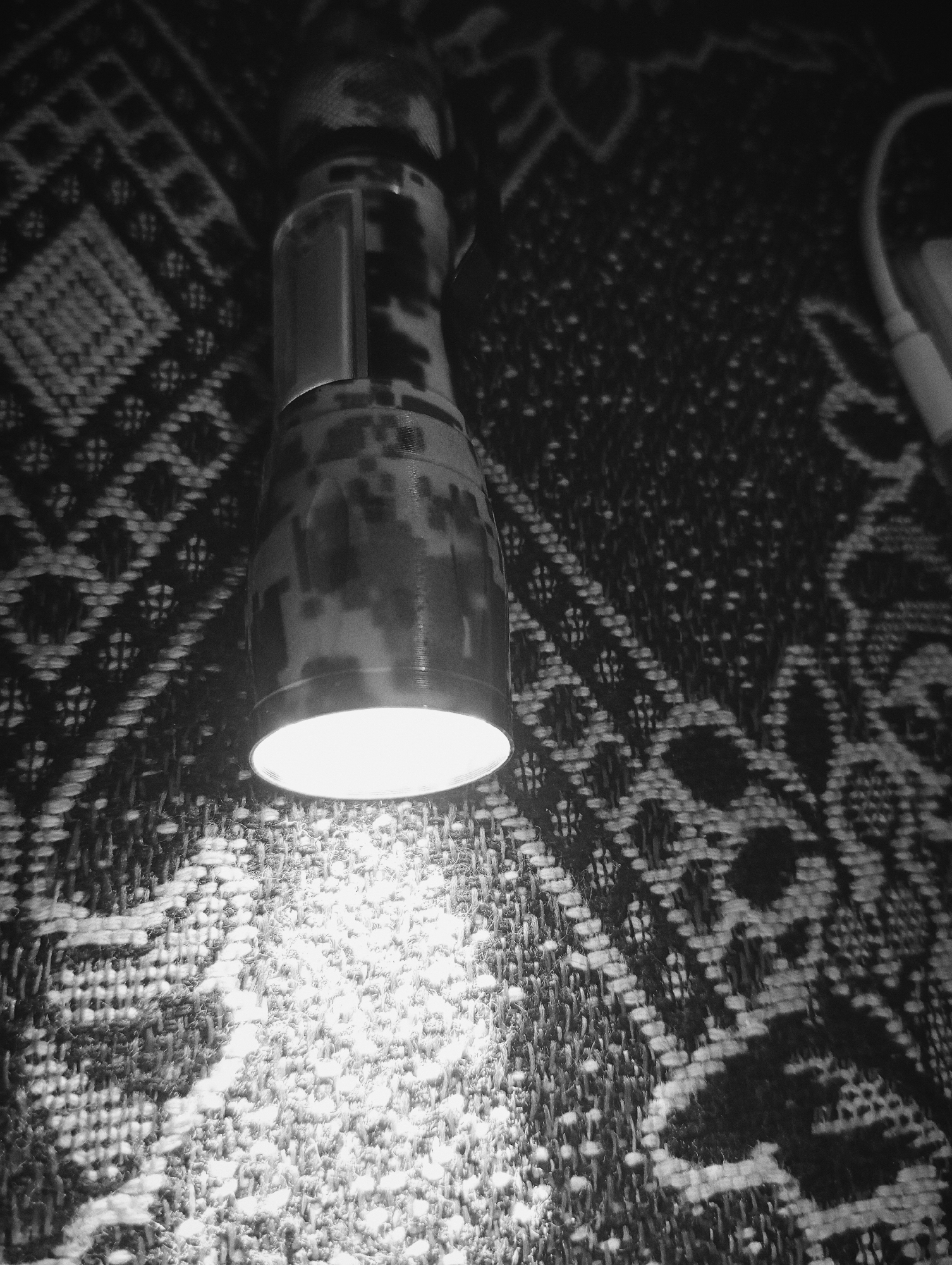 A black and white image shows a flashlight illuminating a patterned surface. The flashlight, with a mottled design, is positioned near the top of the frame, casting a bright circular light onto the textured fabric below. The surrounding area is darker, allowing the illuminated section to be clearly visible. A cable is visible in the background.
The location in the city of Lassan, Pakistan is not identifiable based on the information of the image.