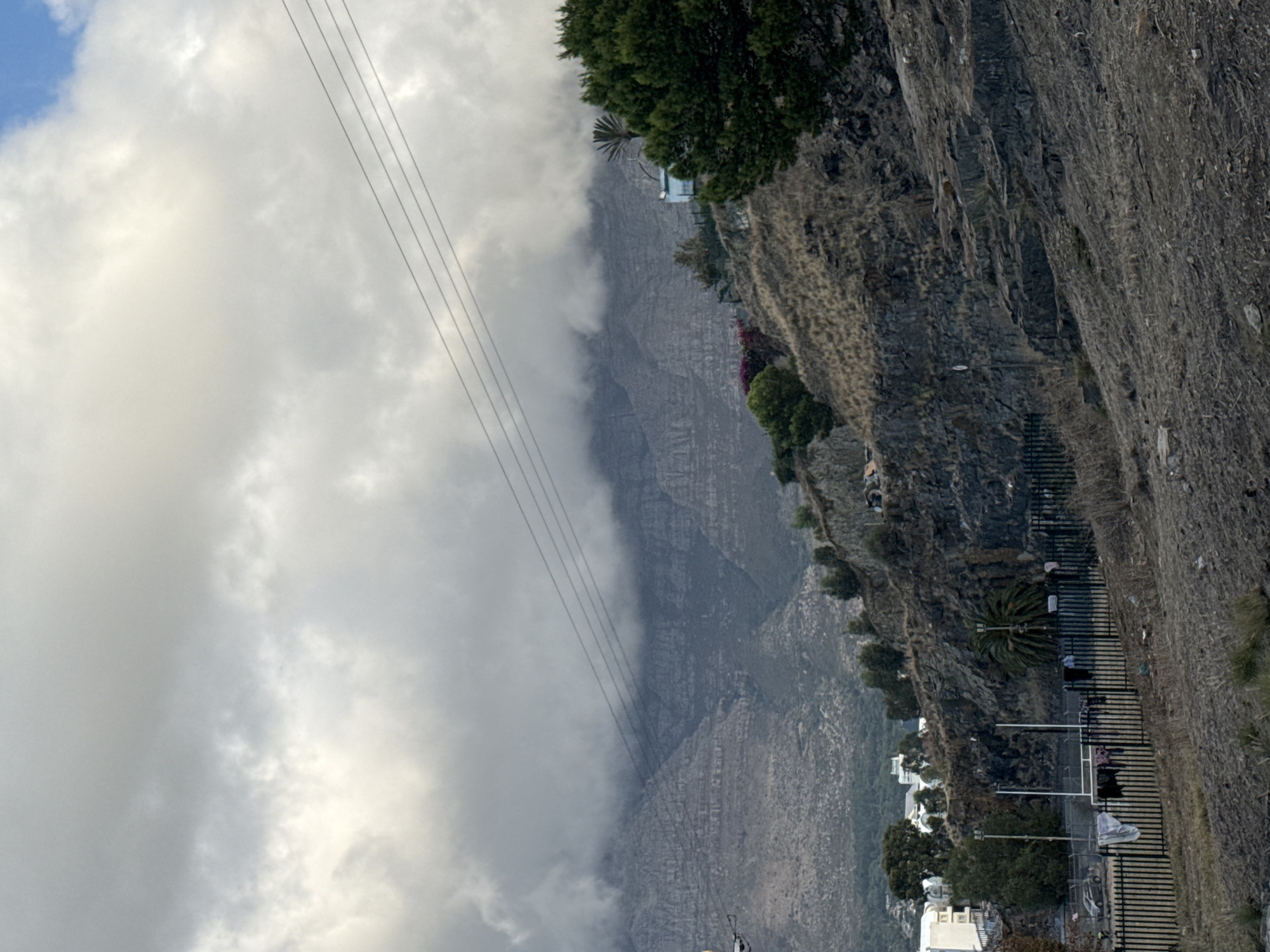 The image captures a view of Table Mountain in Cape Town, South Africa. The mountain is partially obscured by clouds, with power lines visible in the foreground. The right side of the image features a steep, rocky cliffside with vegetation and a fence. A road with parked cars is visible below the cliff. Buildings are nestled among the trees and at the base of the mountain.