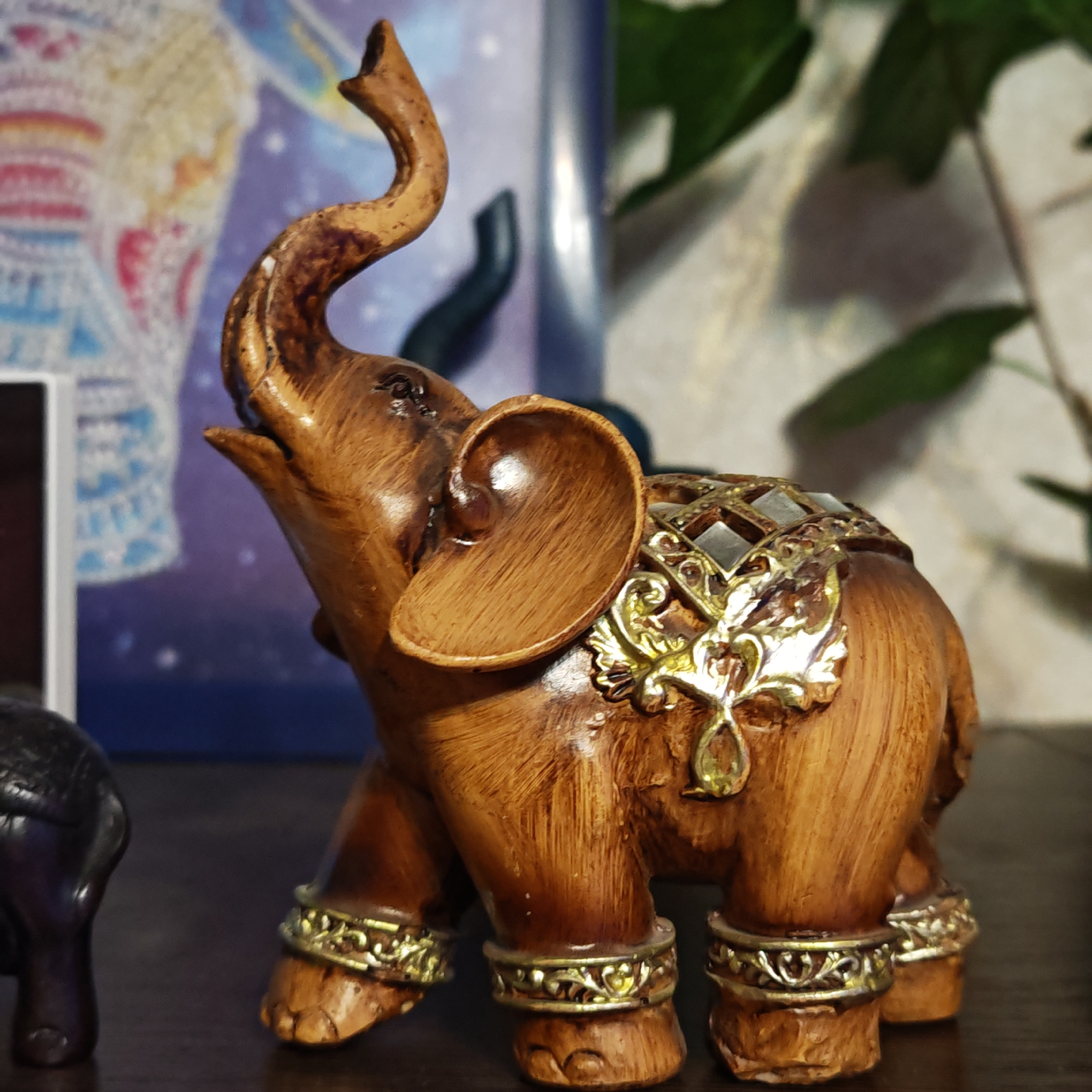 A figurine of an elephant, decorated with gold accents and standing on a dark surface, is the central focus of the image. A blurry blue picture and foliage are in the background. The image was taken in an indoor setting, but there is no information about Rostov, Russia.