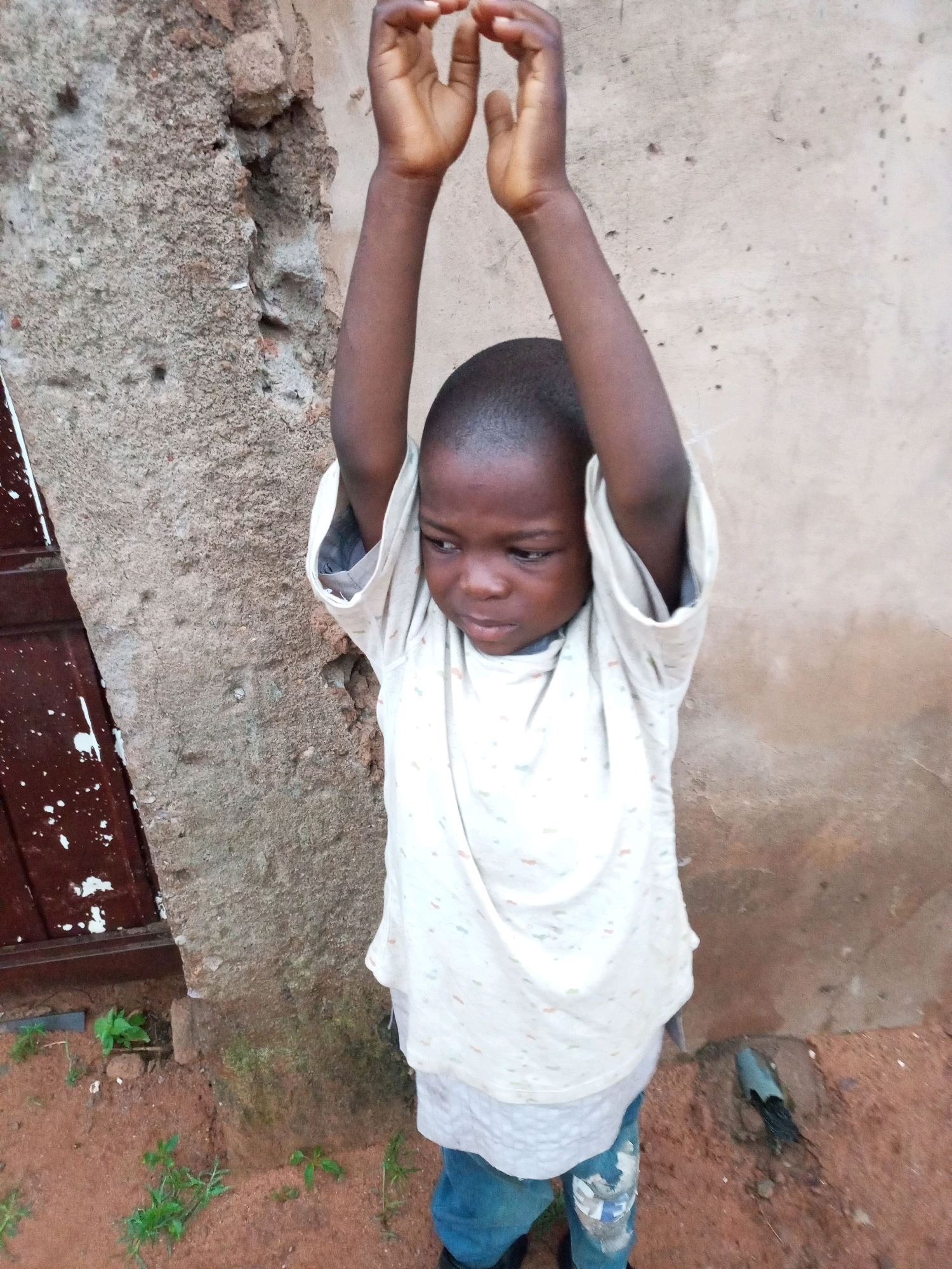 A young boy stands in front of a rough concrete wall in a dirt area. He is wearing a light-colored t-shirt with small colorful specks and blue jeans. His arms are raised above his head, and his head is slightly bowed, with his gaze directed downwards. The wall appears weathered and textured, with a dark brown door or panel to the left of the boy. The ground is covered in reddish-brown dirt with sparse green vegetation growing in patches. A dark pipe or drain is visible in the dirt to the right of the boy. The lighting suggests it is daytime, with natural light casting shadows. This scene is located in Tempure, Nigeria.