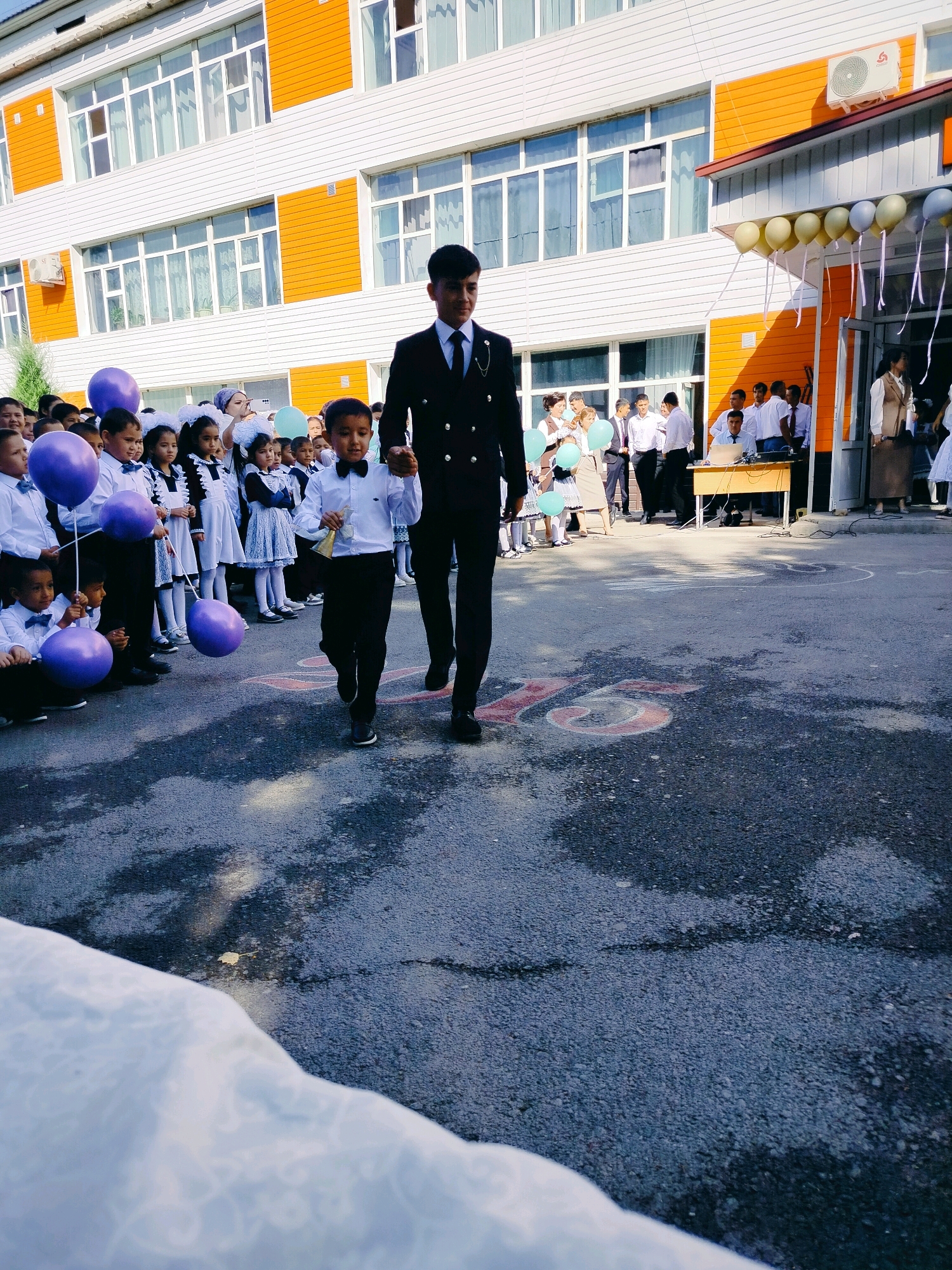 In the city of Kölkent, Kazakhstan, this image captures a school event outside an orange and white building. Two young boys are walking side-by-side, with the taller boy, dressed in a dark suit and tie, holding the hand of the smaller boy, who is wearing a white collared shirt, black bow tie, and black pants. The smaller boy is carrying a small bell-shaped object in his right hand. Behind them, a crowd of children, mostly girls in white dresses and dark uniforms with white bows, are lined up, many holding purple balloons. Adults are also present in the background, some near the entrance of the building where balloons are strung across the awning. The ground is asphalt, with chalk markings of the number "75" visible in red. The scene suggests a festive occasion, possibly the start of the school year or a special celebration.
