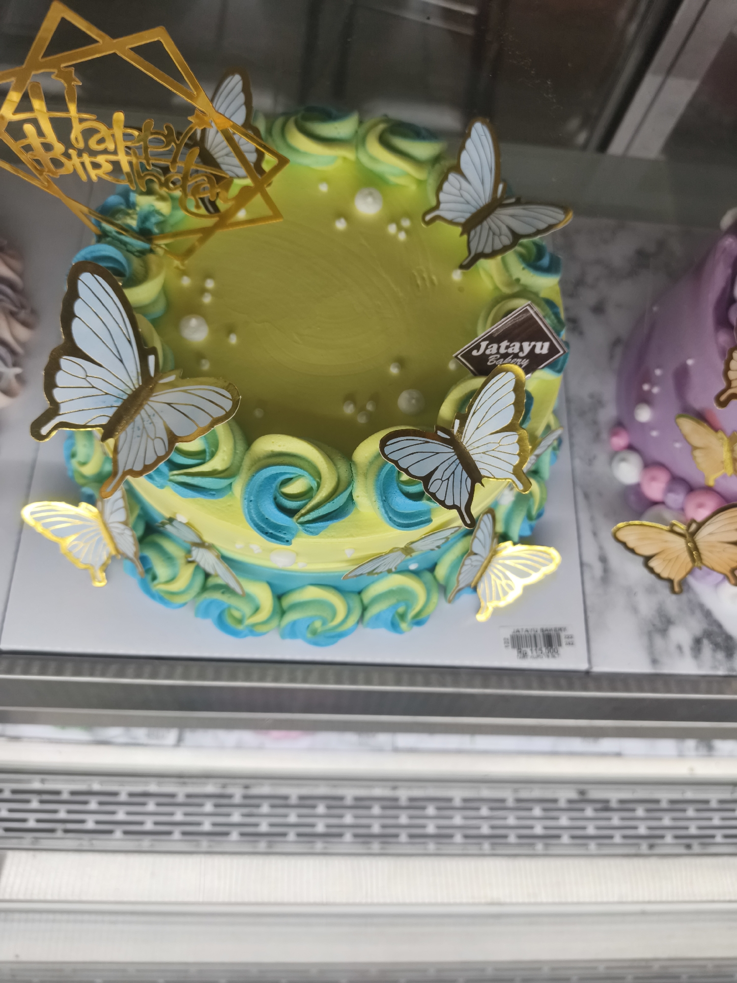 A close-up, eye-level shot displays a yellow and blue swirled cake adorned with white and gold butterflies. A gold "Happy Birthday" topper is placed on the upper left of the cake. A brown rectangular sticker with "Jatayu Bakery" is positioned on the right side of the cake. The cake is situated on a white surface, likely a display in a bakery. In the background, another purple cake with colorful decorations is partially visible. The overall setting appears to be an indoor environment with display shelving and lighting.