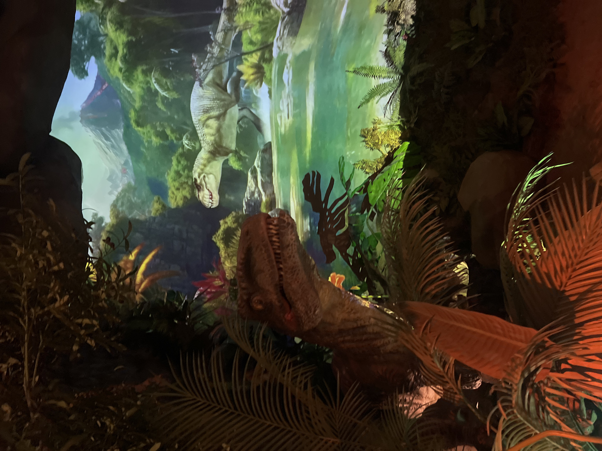The image shows a diorama or exhibit featuring two dinosaurs. In the foreground, to the left, a detailed model of a raptor is visible, partially obscured by lush, dark green foliage and reddish-orange leaves. The raptor has its head turned to the right, revealing sharp teeth and red markings on its face. Behind the raptor, a large, projected image depicts a prehistoric landscape. This background scene includes a vibrant green body of water, trees, rocks, and a distant volcano with lava flowing down its side. Another dinosaur, a Tyrannosaurus Rex, is depicted mid-leap in the projected background, appearing to run across the landscape. The lighting in the foreground is dim and warm, with some areas highlighted by a reddish glow, likely from artificial lights illuminating the foliage and the raptor model. The overall impression is a simulated prehistoric environment designed for visual display, possibly in a museum or theme park. The location is not specified beyond "Unknown, Unknown."