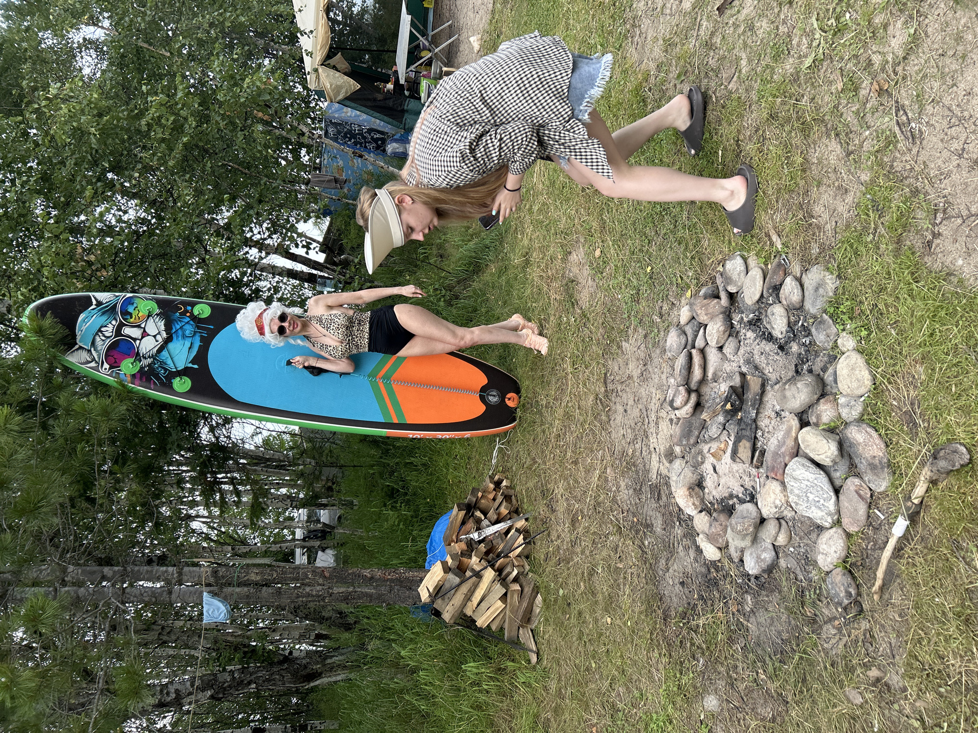 The image depicts an outdoor scene with two individuals in a grassy area, likely a campsite, near a body of water. The location is identified as Tolbazikha, Russia.
In the foreground, a campfire pit is visible, surrounded by stones and ash. A small pile of chopped firewood is stacked to the left of the pit, with a tool resembling a saw leaning against it.
One person, identified as a woman, stands to the left of the frame, leaning against a large, colorful stand-up paddleboard. She is wearing a leopard-print swimsuit, a white wig, sunglasses, and light-colored shoes.
The second person, also a woman, is positioned in the middle ground to the right, facing away from the viewer. She is wearing a plaid jacket over a denim skirt and dark slides. She is looking down at a mobile phone in her hands.
In the background, trees and vegetation are abundant, with a glimpse of a tent and a portable table set up in a camping area. A blue garment is hanging from a clothesline strung between two trees. The overall environment suggests a natural, recreational setting.