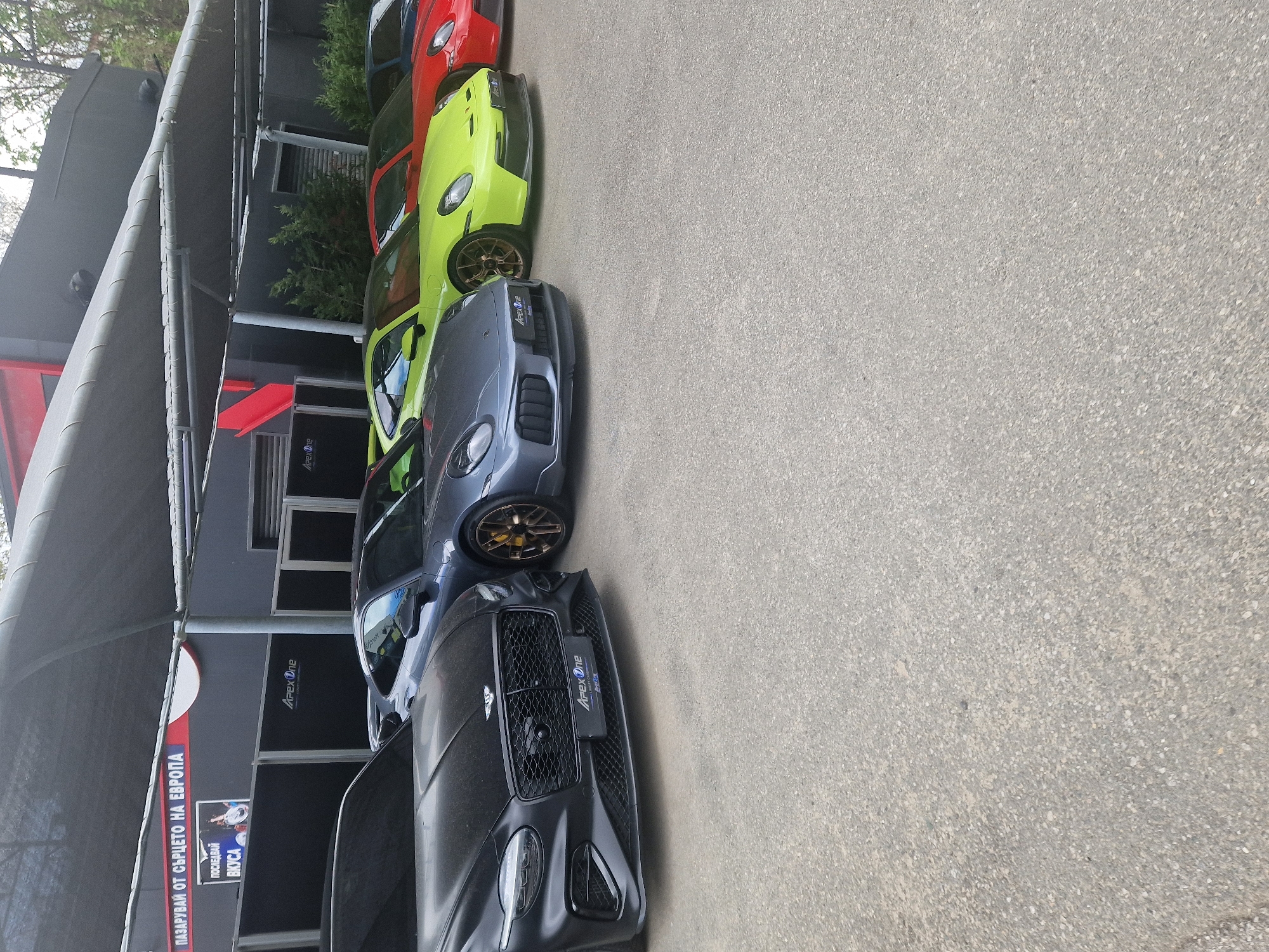 An outdoor, rotated eye-level shot captures a line of luxury sports cars parked on asphalt next to a building under bright daylight. The ground is grey tarmac with visible aggregate.
In the foreground, a dark grey or black Bentley Continental GT is prominently featured, displaying a custom front license plate holder with the "Apex One" logo. Behind it, a dark grey Porsche 911, possibly a GT3, is visible, sporting gold/bronze multi-spoke wheels and an "Apex One" sticker on its front bumper. Following this, a vibrant lime green Porsche 911, also with gold/bronze wheels and an "Apex One" sticker, is parked. Further down the line, a red sports car, likely another Porsche, is partially visible, with a small section of a blue car at the very top of the image.
The cars are parked alongside a dark-colored building with windows, partially covered by a large, grey canopy structure. The building also displays the "Apex One" logo. On the grey canopy, a sign in Cyrillic reads: "ПАЗАРУВАЙ ОТ СЪРЦЕТО НА ЕВРОПА" (Pazaruvay ot surtseto na Evropa, meaning "Shop from the heart of Europe") and below it, "ПОСЛЕДНИ ВКУСА" (Posledni Vkusa, meaning "Latest Taste" or "Last Taste"). Greenery and trees are visible beyond the building structure. The clear lighting suggests it is daytime, likely a sunny morning or afternoon. No people are visible. The scene suggests a high-end automotive facility, such as a dealership, detailing shop, or tuning garage.