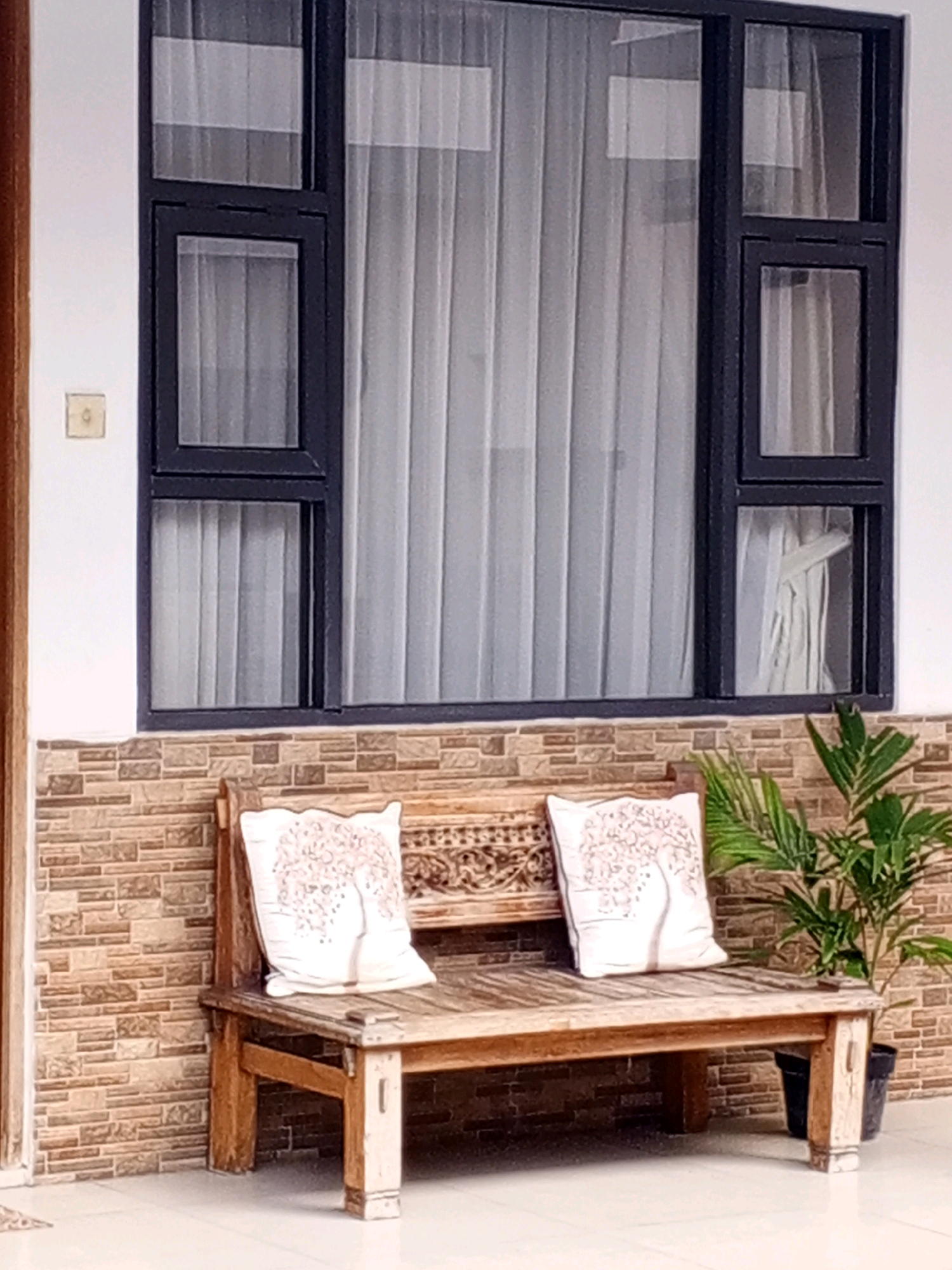 An outdoor scene in Denpasar, Indonesia, features a wooden bench with two decorative pillows in front of a window. The bench is made of light-colored wood and has intricate carvings on its backrest. The pillows are white with a pink tree-like pattern. To the right of the bench, a potted green plant with palm-like fronds sits on the tiled floor. The window behind the bench has a dark frame and sheer white curtains. The wall below the window is covered with a brick-patterned tile. The floor is a light-colored tile.