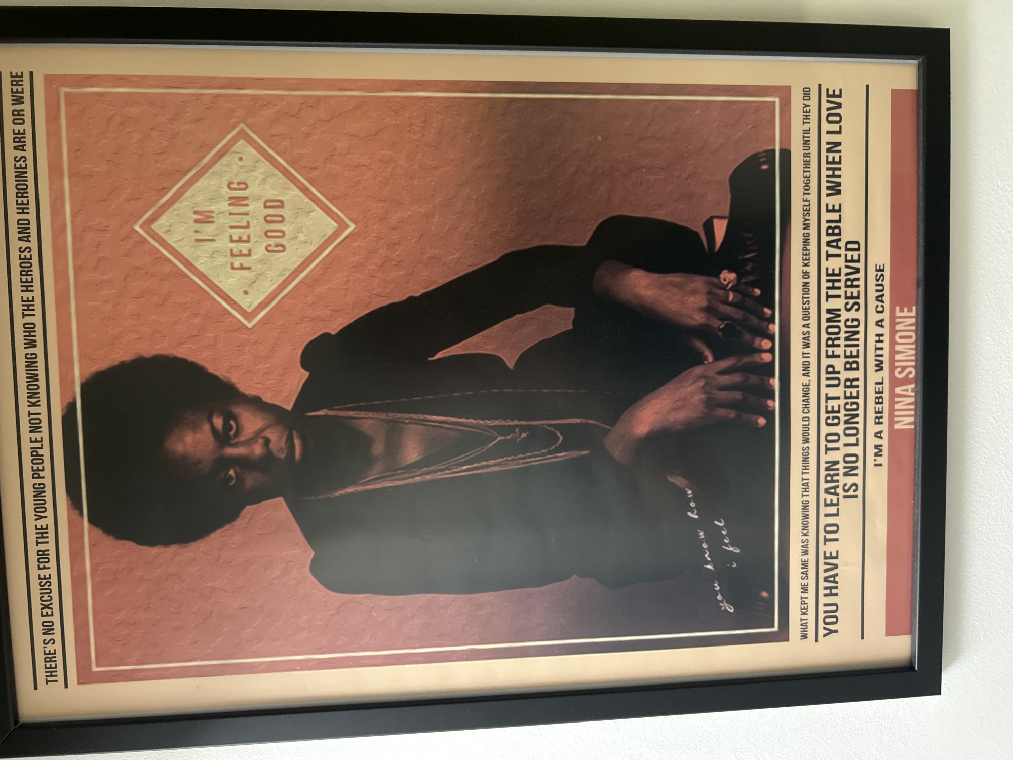 A framed poster of Nina Simone is hanging on a white wall. The poster features a close-up portrait of Simone with her signature afro hairstyle. She is looking directly at the camera with a serious expression. The poster has text in several places, including a quote that reads, "THERE'S NO EXCUSE FOR THE YOUNG PEOPLE NOT KNOWING WHO THE HEROES AND HEROINES ARE OR WERE". Below her image, there is another quote in handwritten script that says, "you know how i feel". The bottom section of the poster contains more text, including the lines, "WHAT KEPT ME SAME WAS KNOWING THAT THINGS WOULD CHANGE, AND IT WAS A QUESTION OF KEEPING MYSELF TOGETHER UNTIL THEY DID", "YOU HAVE TO LEARN TO GET UP FROM THE TABLE WHEN LOVE IS NO LONGER BEING SERVED", "I'M A REBEL WITH A CAUSE", and "NINA SIMONE". A small diamond shape with the words "I'M FEELING GOOD" is also visible on the poster. The poster is framed in black and hangs on a plain white wall. The location of the poster is not specified in the image, but the user stated it is in Sofia, Bulgaria.