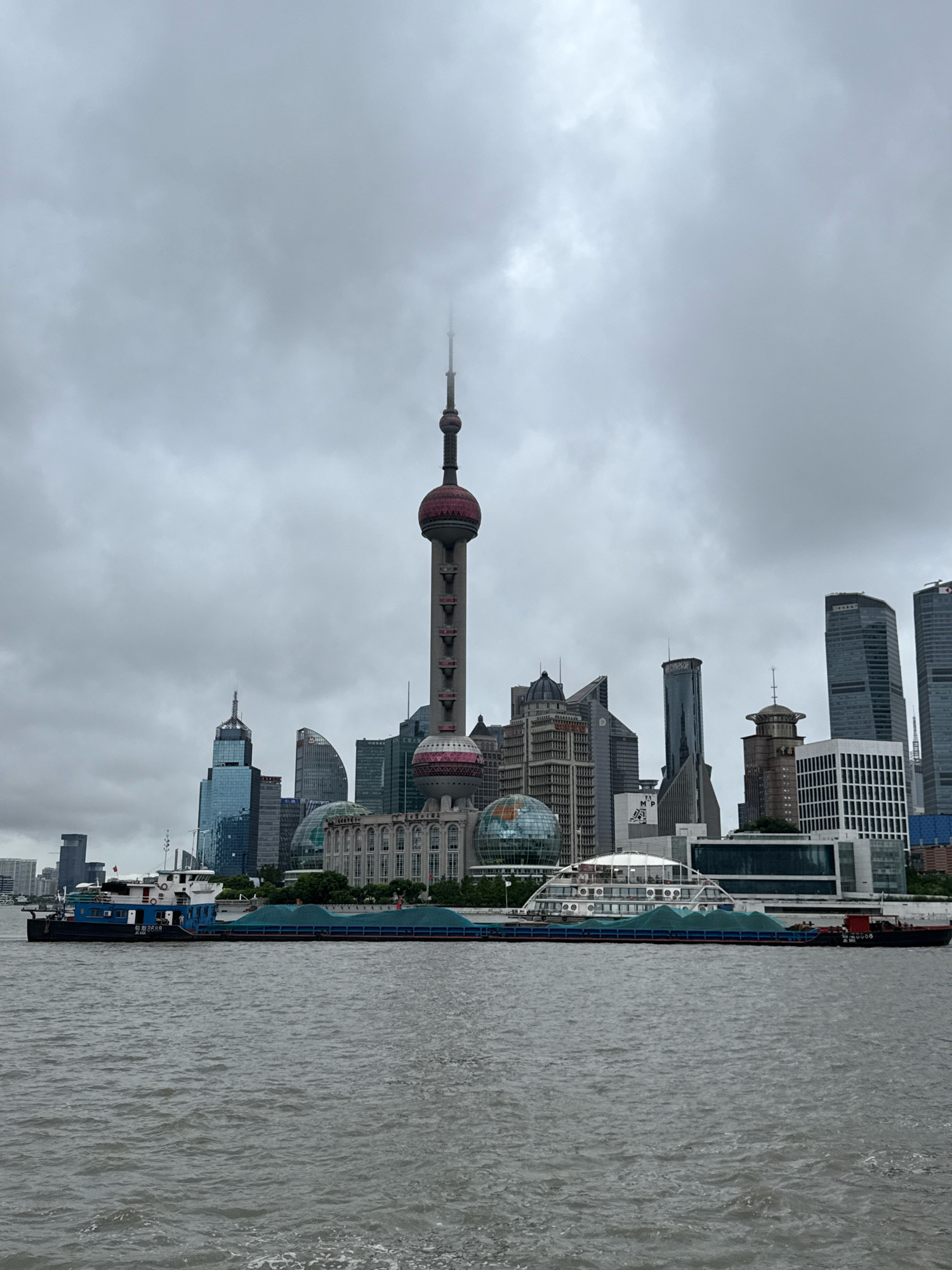This is a full-frame, eye-level, landscape photograph of the Shanghai skyline viewed from the Huangpu River. The sky is overcast and grey, obscuring the tops of some of the taller buildings. In the foreground, the murky, grey-green water of the Huangpu River is visible, with gentle ripples across its surface. A large, dark-colored barge carrying a load of green material, possibly sand or gravel, is positioned horizontally across the middle of the frame, moving from left to right. The barge has a blue section with white lettering and a smaller white superstructure at its rear. Behind the barge, the iconic skyline of Shanghai's Pudong district rises. Dominating the center of the skyline is the Oriental Pearl Tower, a distinctive television tower with a red and white sphere near its top and a smaller sphere lower down. To the left and right of the tower are numerous modern skyscrapers of varying heights and architectural styles, including a building with a distinctive blue facade and pointed spire, and another with a curved, glass-like exterior. Several other prominent buildings, including one with a dome-shaped structure and another that appears to be a large, white, modern building with a curved roof, are also visible. The overall scene depicts a busy urban waterfront under a cloudy sky.