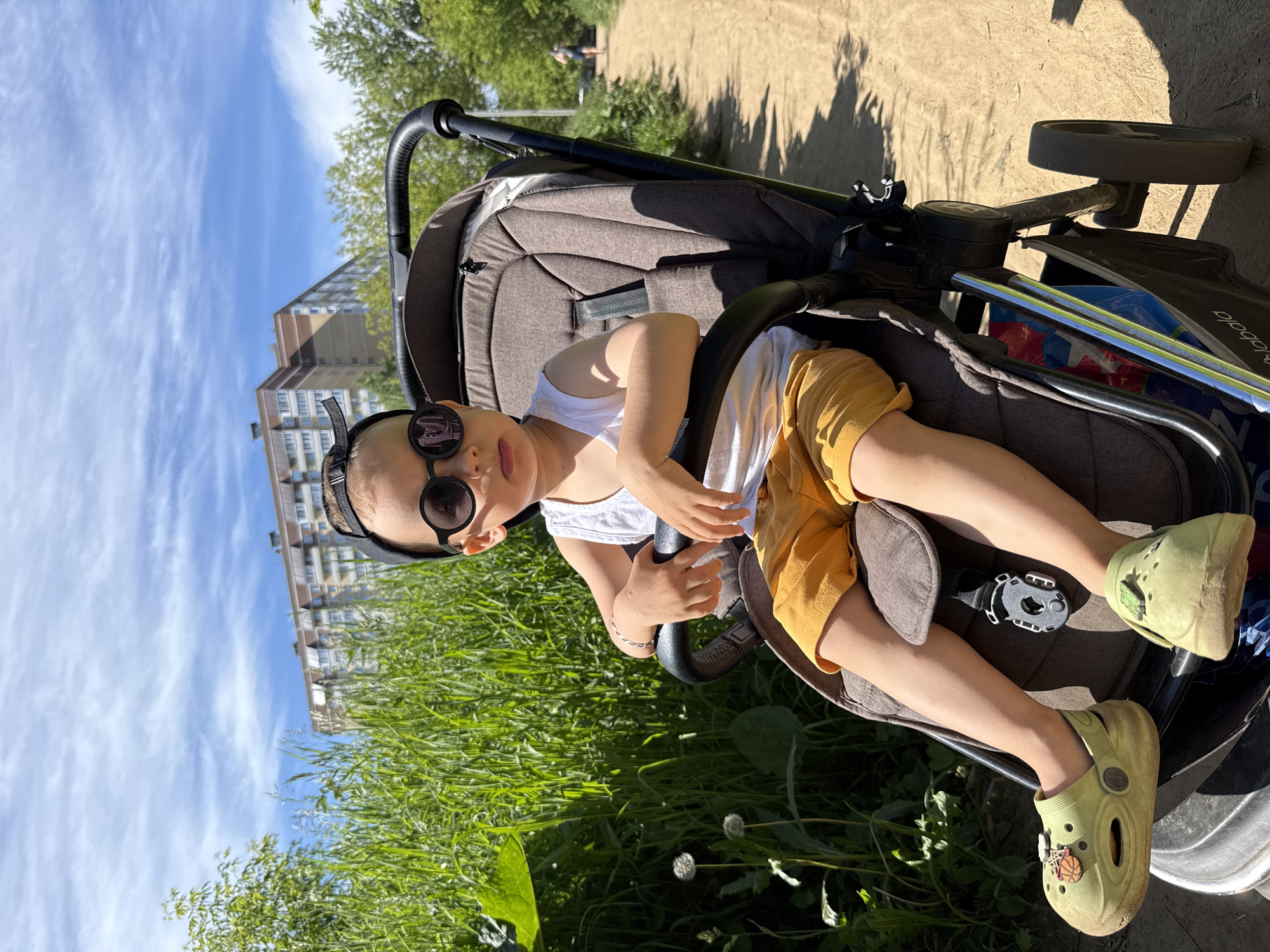 A young child is seated in a stroller, looking directly at the camera. The child is wearing a white tank top, yellow shorts, black sunglasses, and light green clogs. The stroller is dark brown with a gray seat. In the background, there is a tall, light-colored apartment building, lush green grass, and trees. The sky is blue with scattered white clouds. The sun is shining brightly, casting shadows on the ground, indicating it is daytime. The scene appears to be outdoors, possibly in a park or residential area. The child's expression is neutral. There is no visible text in the image. The overall impression is a sunny day outing with a child in a stroller.