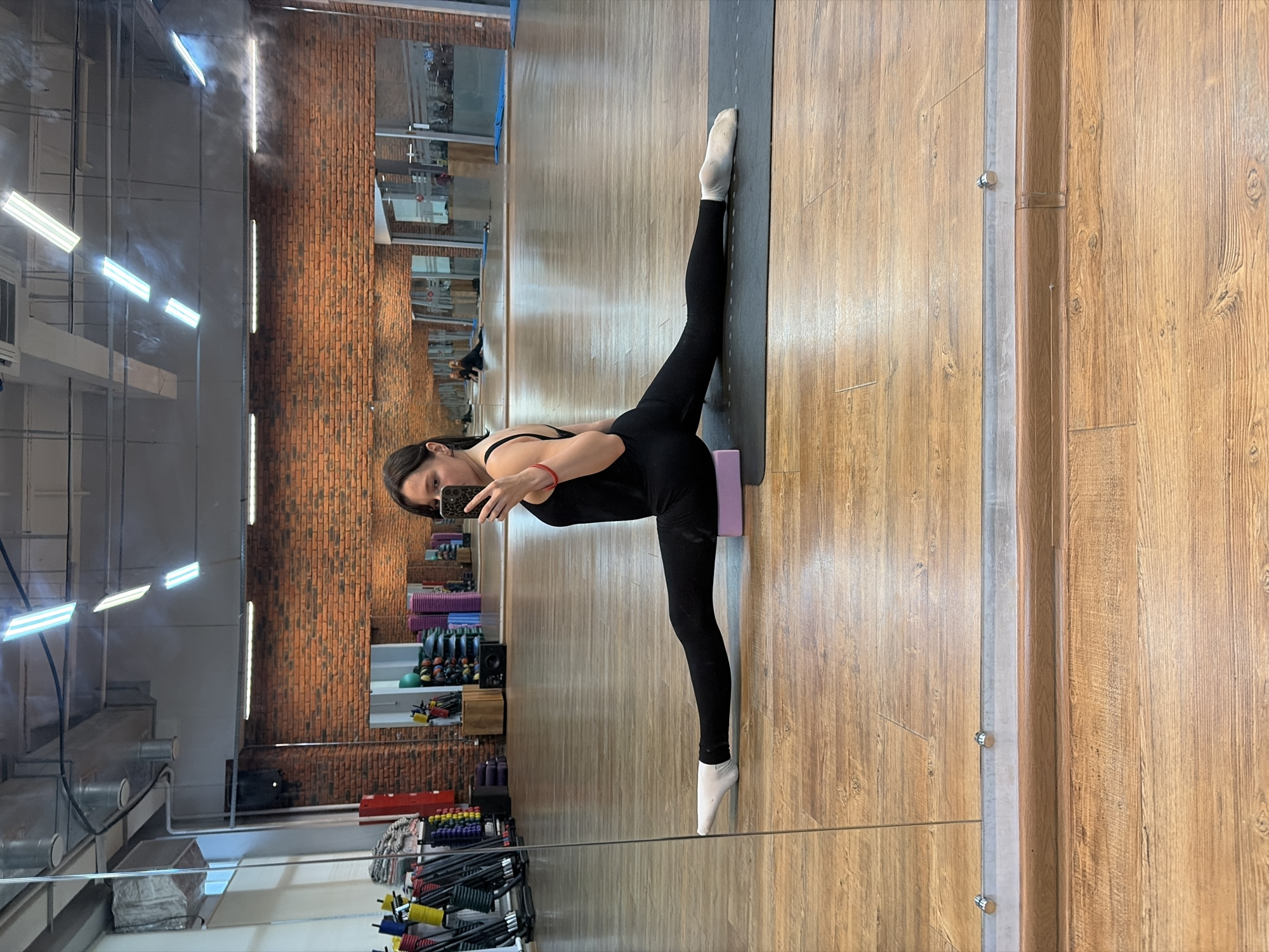 A woman is captured in a full-body, vertical shot, performing a split on a yoga mat in what appears to be a gym or dance studio. She is wearing a black leotard and black leggings with white socks. Her left leg is extended straight up in the air, with her foot on the mat, while her right leg is bent at the knee and resting on a pink yoga block. Her body is angled, and she is holding a phone in her right hand, taking a selfie. The background reveals a large mirror reflecting the studio, showing brick walls, fluorescent lighting, and various gym equipment such as weights, yoga blocks, and resistance bands neatly organized on shelves. The flooring is light-colored wood. The lighting suggests it is daytime, but the interior setting makes it difficult to determine the exact time or weather conditions. The woman's expression is neutral, focused on her pose. The setting, with its gym equipment and mirrors, indicates a place for physical training. The reflection in the mirror shows more of the studio space, including what looks like a reception area and more workout equipment.