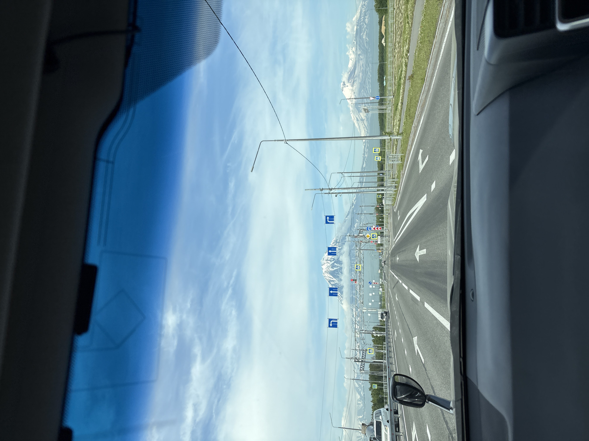 The image is taken from the interior of a vehicle, looking out through the windshield. The view is of a multi-lane highway with traffic signs and streetlights lining the sides. In the distance, snow-capped mountains are visible under a partly cloudy sky. The road has white lane markings and several vehicles are on the highway. One vehicle, a white SUV, is in the left lane. Traffic lights and directional signs are also present, including signs indicating left turns and pedestrian crossings. The foreground shows the dashboard of the car and a rearview mirror. The scene appears to be in Yelizovo, Russia, given the presence of distinct volcanic mountains in the background.