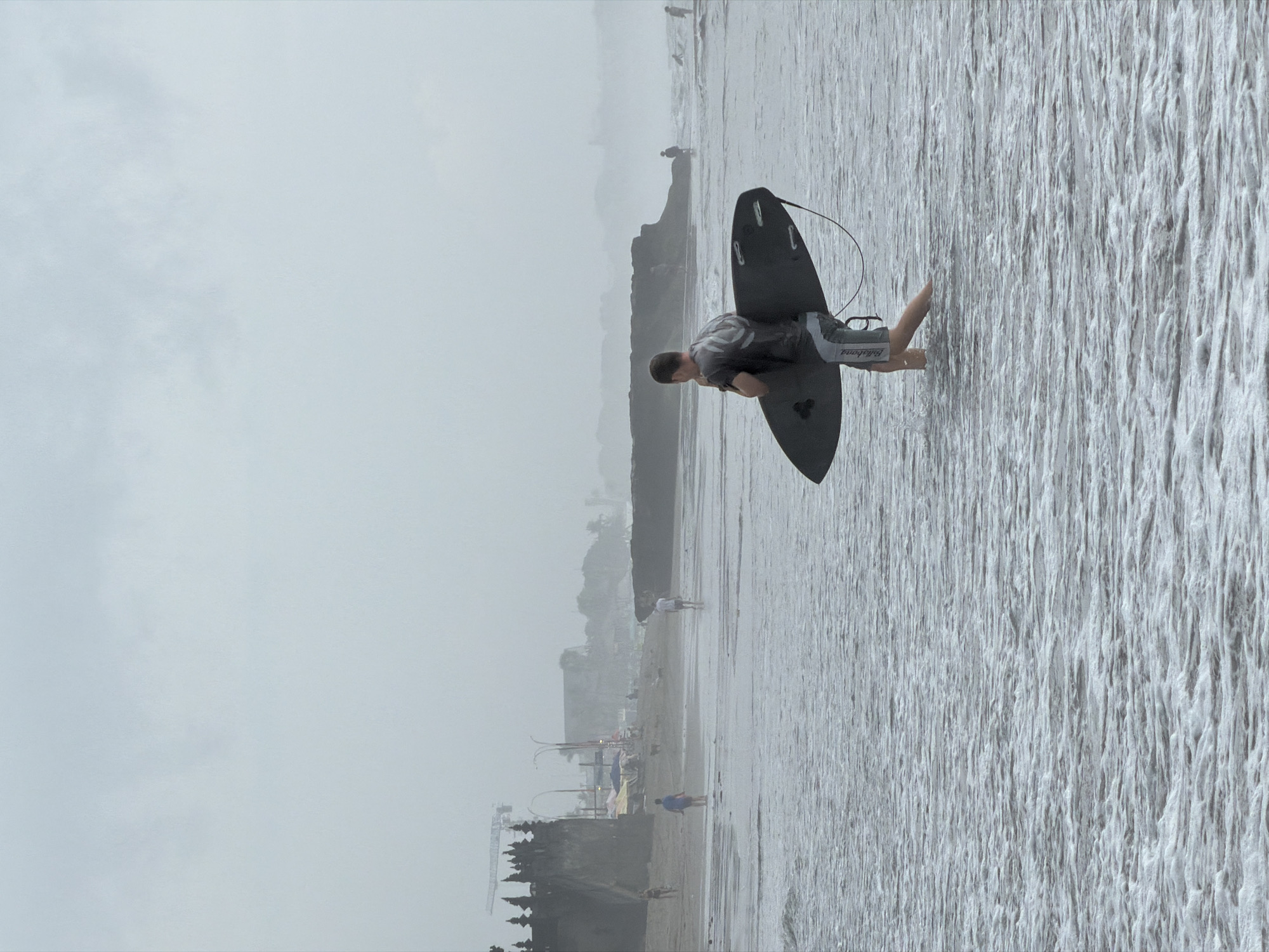 In North Kuta, Indonesia, a man walks into the ocean carrying a surfboard. He is walking towards the left of the frame, away from the shore. The surfboard is black with white fins and is held horizontally across his body. He is wearing grey and white patterned board shorts and a grey t-shirt with a graphic design. The ocean is murky and appears to have a foamy texture from small waves. In the background, the beach is visible with several other people walking and standing. Structures resembling temples or buildings are visible on the shore, along with a construction crane in the distance. The sky is overcast and the overall atmosphere of the image is hazy.