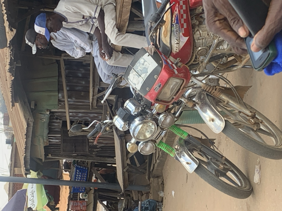 In Egbedi, Nigeria, a daylight scene depicts a street or market environment. Two motorcycles are parked prominently in the foreground on a dirt ground surface. The motorcycle on the left features chrome triple headlights, a chrome front fender, and green covers on its front shock absorbers. The red motorcycle on the right has a square headlight, a small windshield, and "DAYLONG" branding with a red, white, and blue checkered pattern on its fuel tank.
In the mid-ground, two men are seated on a wooden bench, facing right. The man on the left wears a light grey top and trousers with a black and white baseball cap; his arms are crossed. The man on the right wears a light-colored, possibly patterned, tunic and trousers with a blue and white baseball cap; he holds a small, light-colored object in his hands.
Behind the men and motorcycles, a rustic structure composed of wooden planks and corrugated metal sheets, with a rusty corrugated metal roof, functions as a stall or shop. Wooden tables are visible in front of this structure. To the far left, a green and white umbrella branded "Glo Cafe" partially covers a blue plastic crate and other items, suggesting another commercial point. In the immediate foreground, the dark-skinned hands of a person are visible, operating a black and green feature phone resting on a blue cloth.