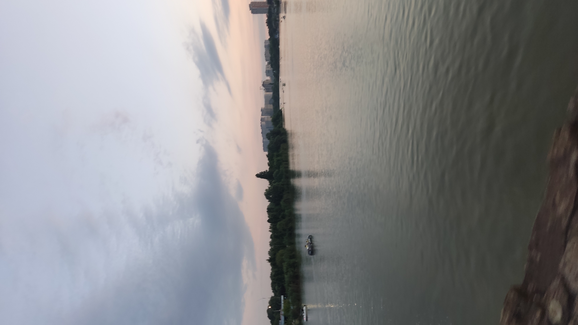 The image depicts a wide, calm body of water, likely a river or lake, under a soft, overcast sky with hints of pink and orange from what appears to be either dawn or dusk. The water surface has gentle ripples. Along the far bank, a dense line of green trees is visible, with some taller, darker trees interspersed. Behind the trees, a city skyline rises, featuring several modern, multi-story buildings, some of which appear to be residential complexes. A few scattered cranes suggest ongoing construction in the city. On the water, there are several small boats, including what appear to be paddle boats, with a few individuals visible in them. One white boat is further to the left, and a darker boat with occupants is closer to the center. The foreground shows a blurry, dark, textured surface, possibly a wooden railing or walkway, suggesting the vantage point is from a bridge or pier. This scene is consistent with the landscape found along the Kuban River embankment in Krasnodar, Russia, particularly in areas featuring recreational water activities and urban development.