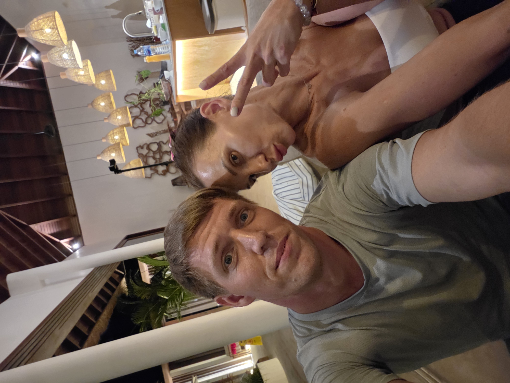 A selfie shot at an angle, possibly with a wide-angle lens. Two individuals are present: A man in the foreground and a woman leaning in behind him. The man is wearing a light green or gray t-shirt. The woman has her hand raised in a peace sign or a victory sign, is wearing bracelets and a white shirt. The location appears to be a resort or upscale accommodation based on the architectural details visible. Features include indoor/outdoor spaces, tropical plants, wooden beams, and modern design elements, with a row of identical lamps. This image was taken in Ubud District, Indonesia.