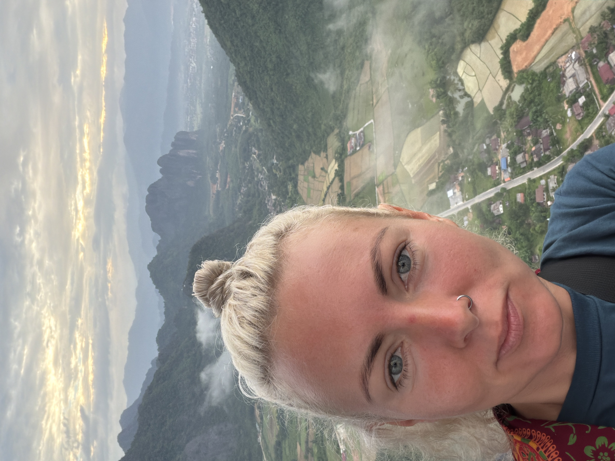 A selfie is taken from a high vantage point overlooking a valley in Hong Ngern Village, Laos. The foreground is dominated by the face of a blonde woman with blue eyes and a nose ring. Her hair is pulled up in a bun. In the background, a lush green valley is visible, with mist clinging to the mountains and a network of rice paddies and scattered buildings. The sky above is cloudy with hints of sunrise.